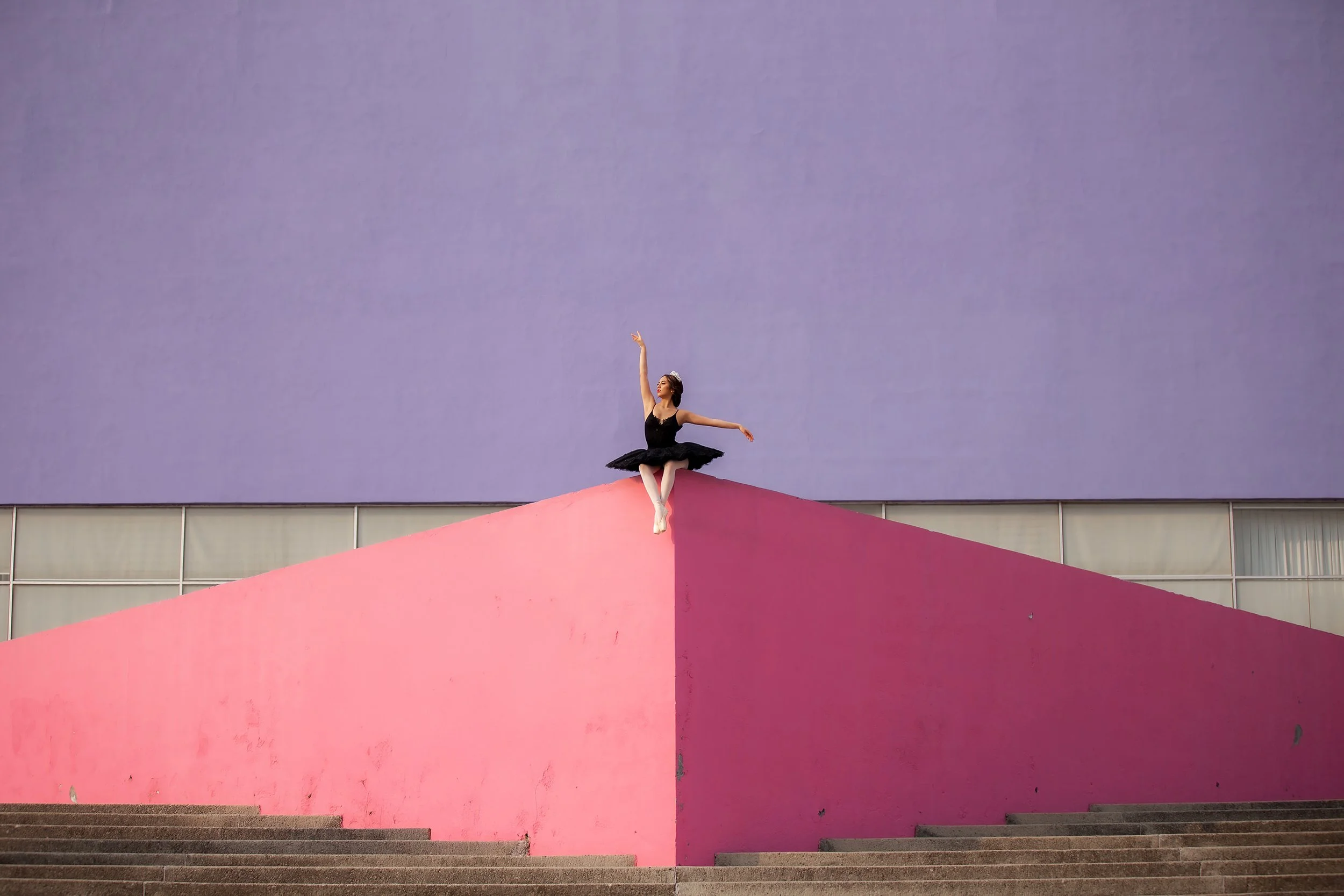 Retrato de mujer sentada sobre estructura geométrica rosa, figura pequeña frente a fondo violeta.
