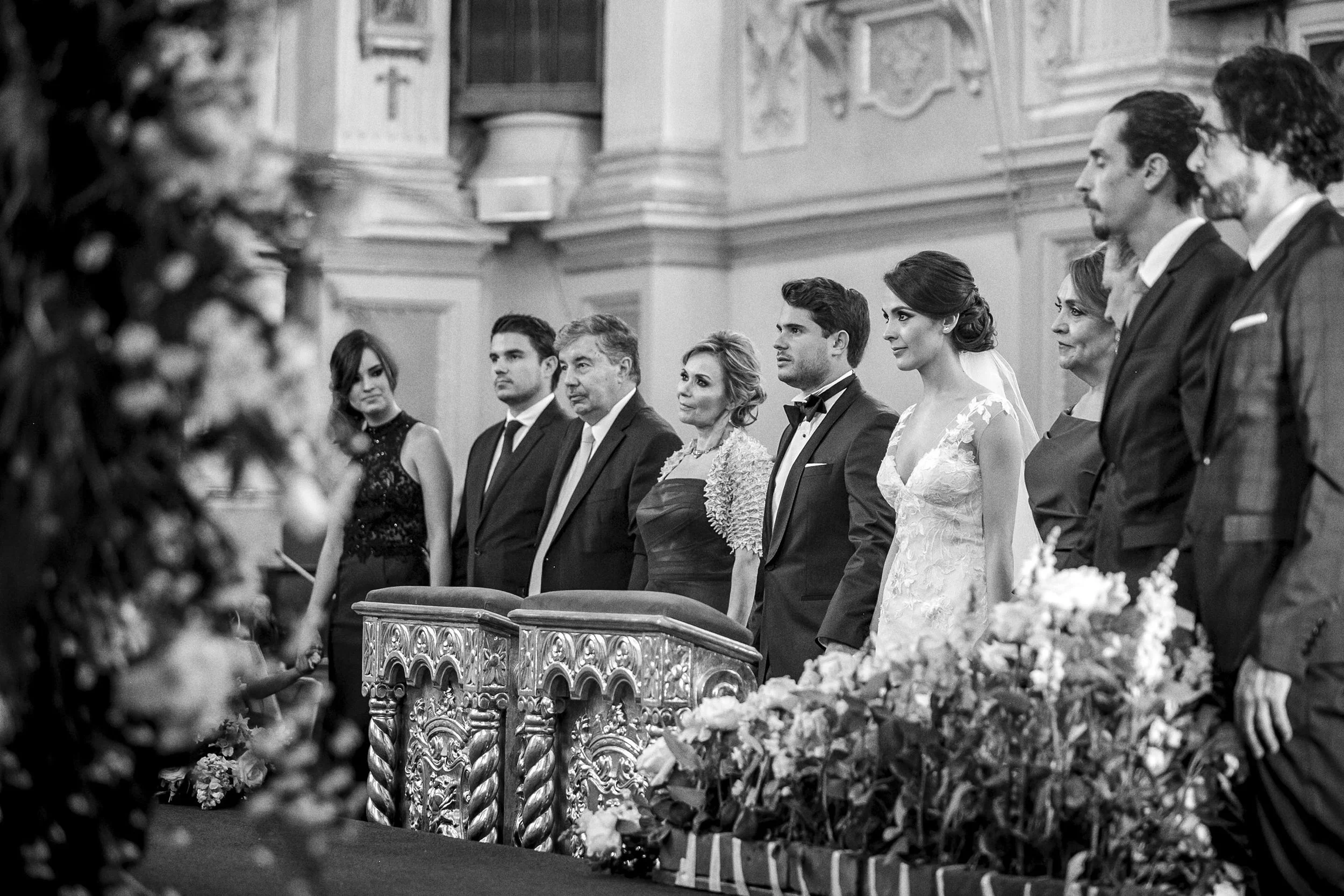 Fotografía en blanco y negro de novios durante la ceremonia, acompañados por familiares en altar.