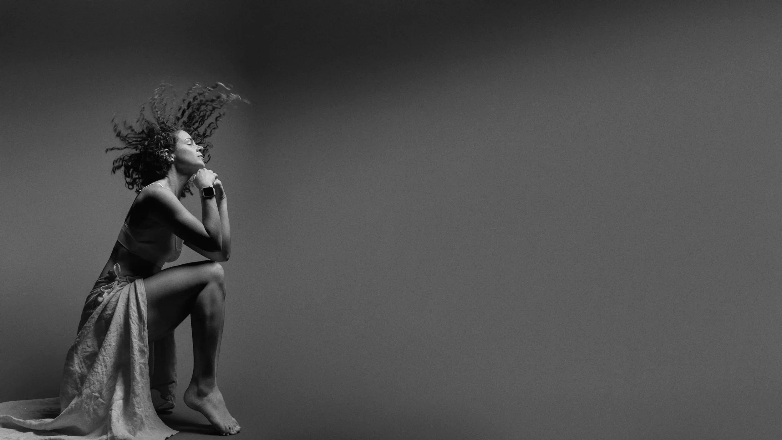 A black and white photo of a woman sitting on a surface, with her hair flowing as if caught in motion, looking contemplative, with her chin resting on her hand. She is wearing light clothing and a smartwatch, and the background is plain.