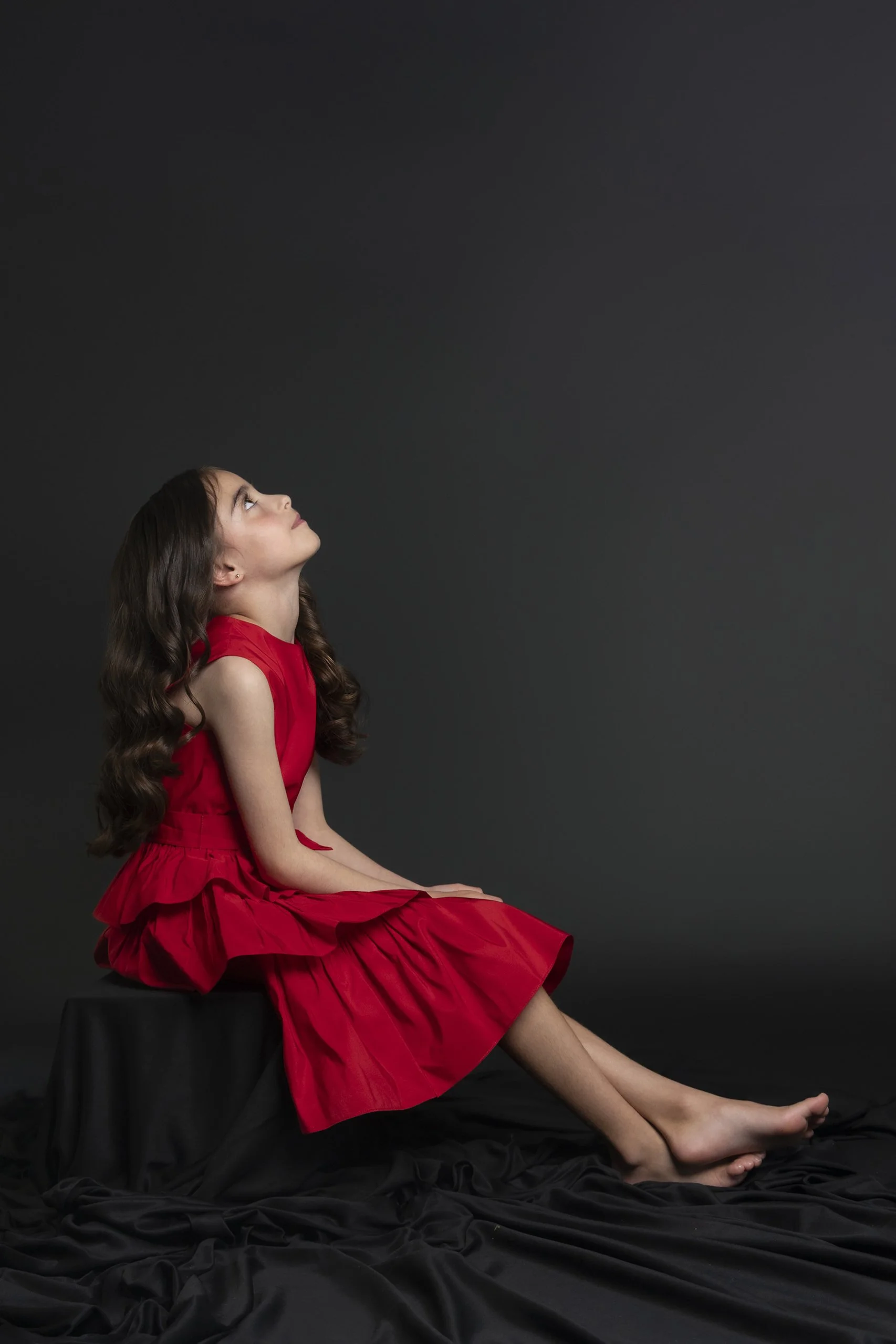 Retrato de niña con vestido rojo sentada, cabeza inclinada hacia atrás y expresión contemplativa en estudio.