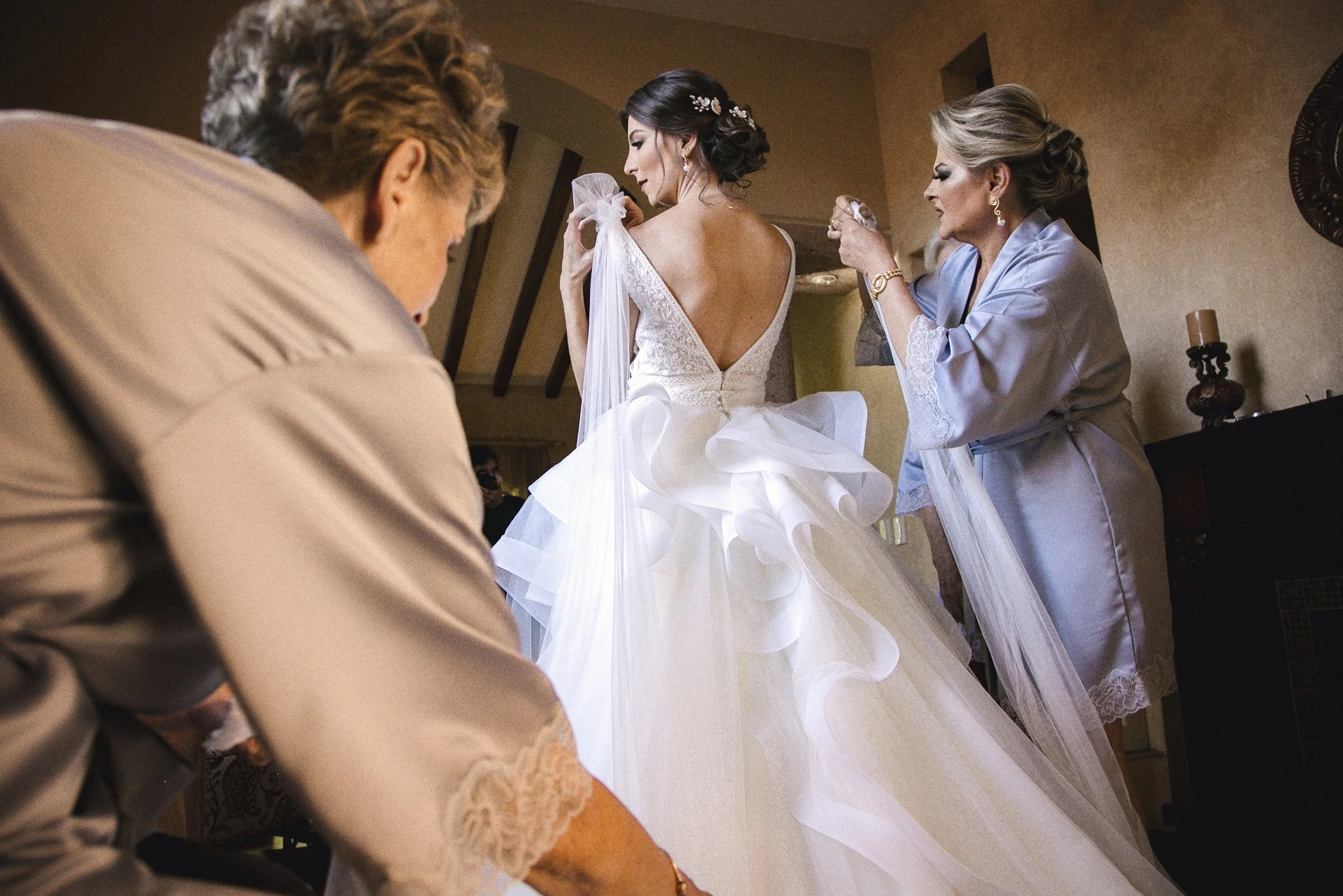 Fotografía documental de novia durante la preparación, asistida por mujeres cercanas, vestido en primer plano.