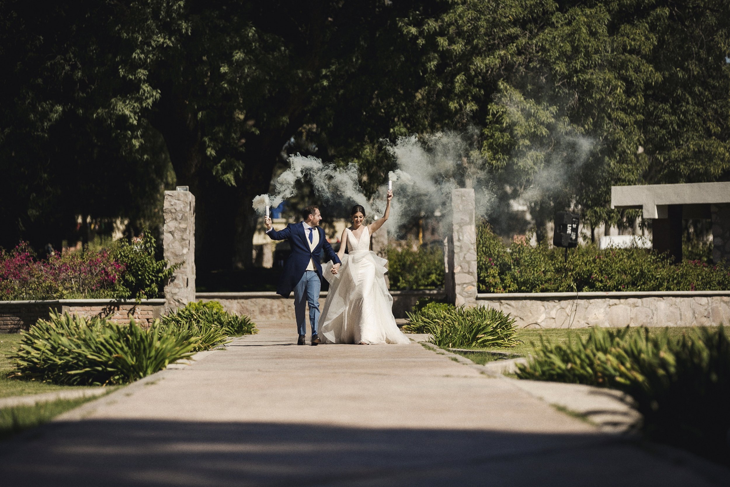 Fotografía documental de novios caminando juntos en jardín después de la ceremonia, acompañados por invitados al fondo.