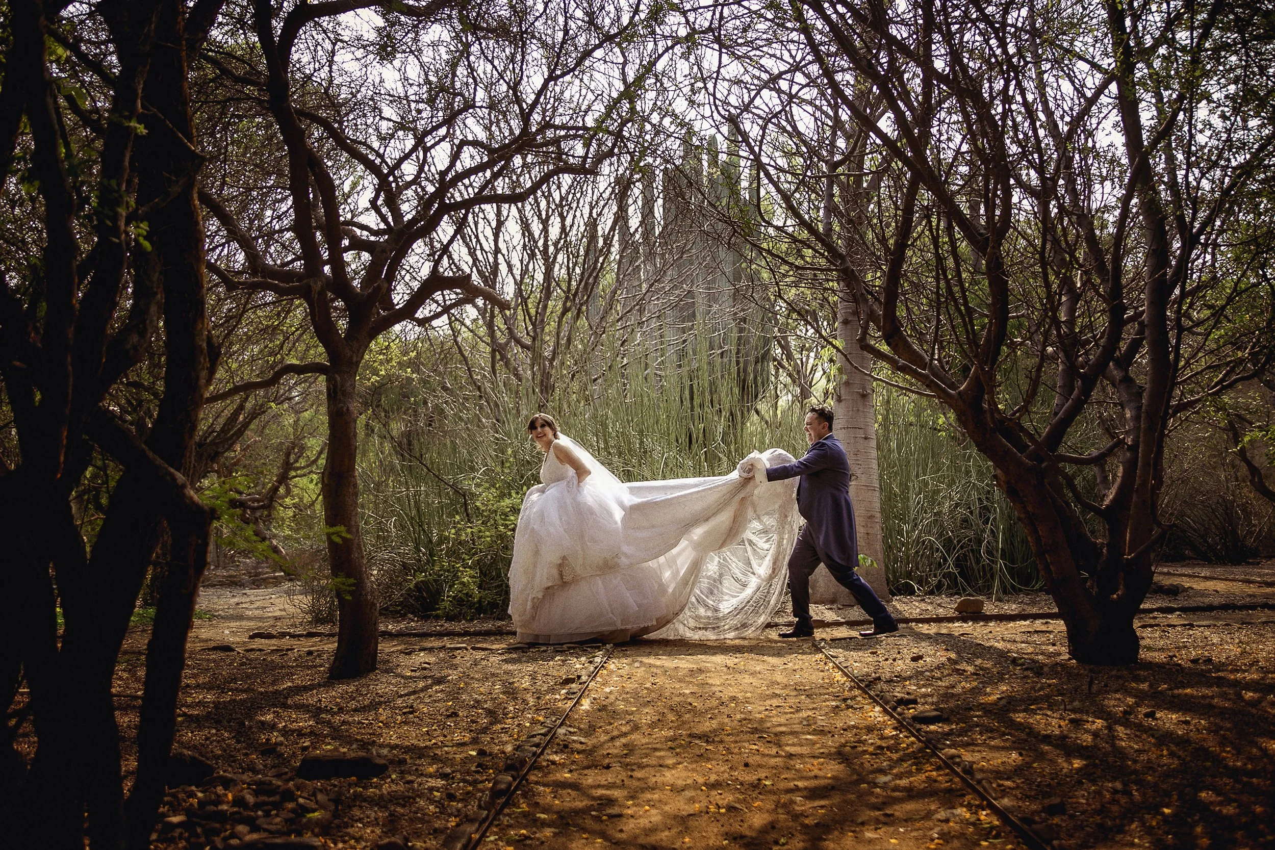 Fotografía documental de novios caminando en exterior arbolado, vestido extendido y movimiento natural.