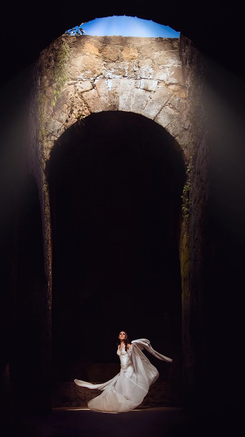 A woman in a flowing white dress standing in a dark, ancient stone structure with an arched opening at the top letting in light.