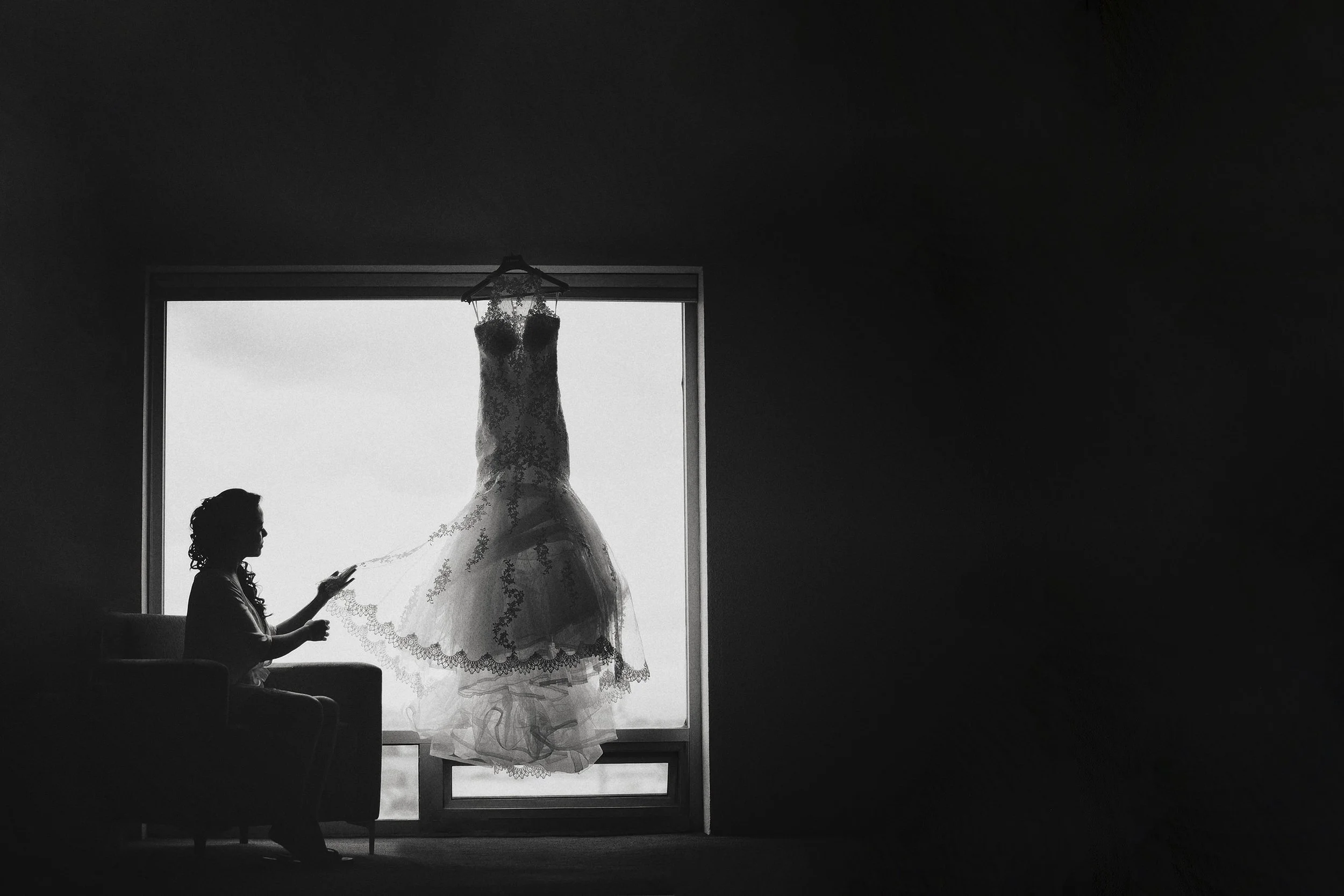 Fotografía en blanco y negro de vestido de novia colgado frente a ventana, luz natural y silueta definida.