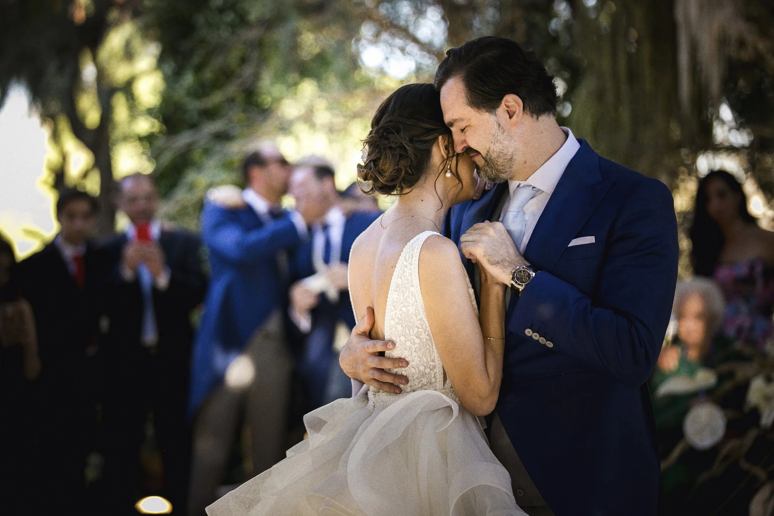 Fotografía documental de novios abrazados durante la celebración, rodeados de invitados desenfocados.