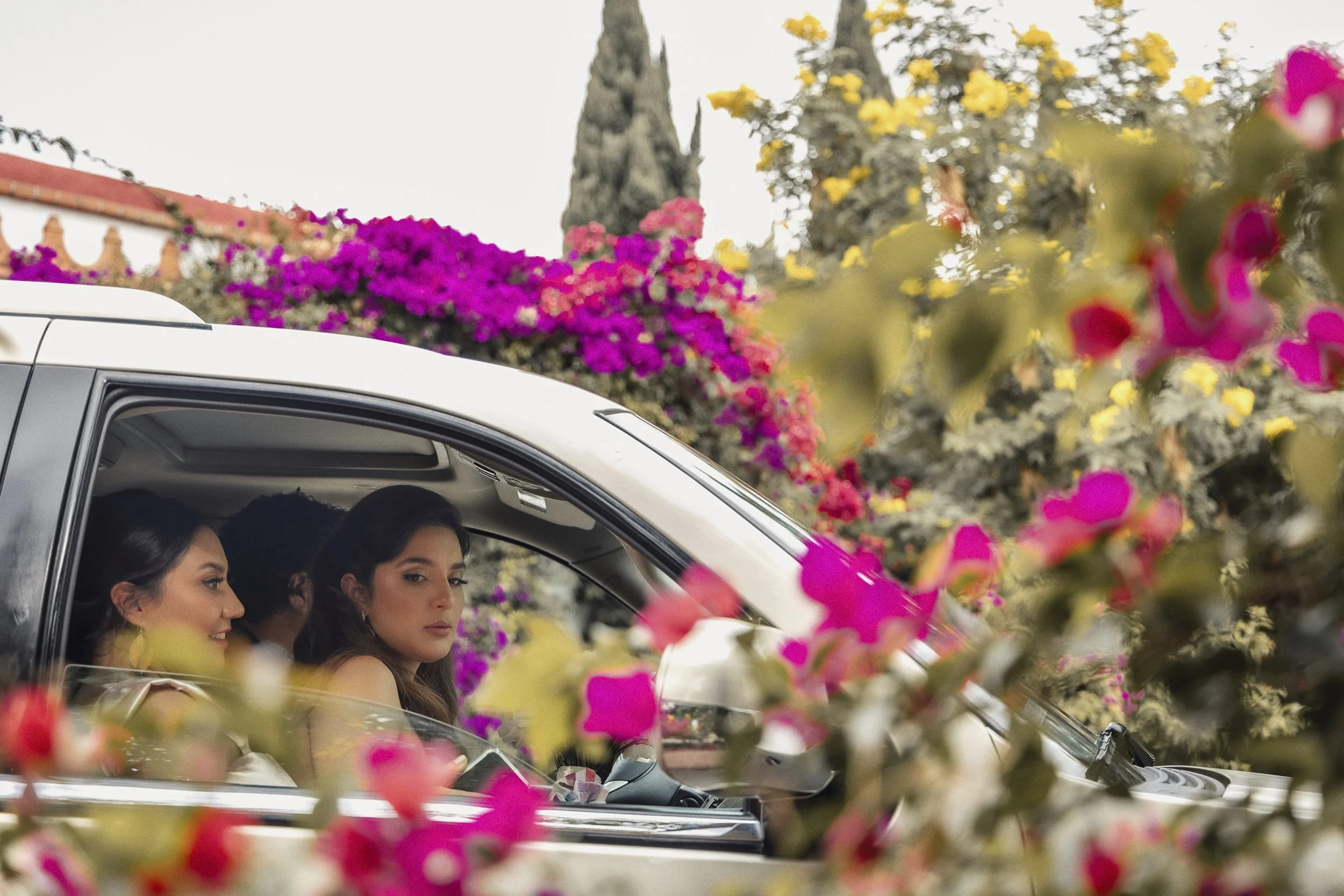Fotografía documental de novia dentro de automóvil rodeado de flores, mirada hacia el exterior.