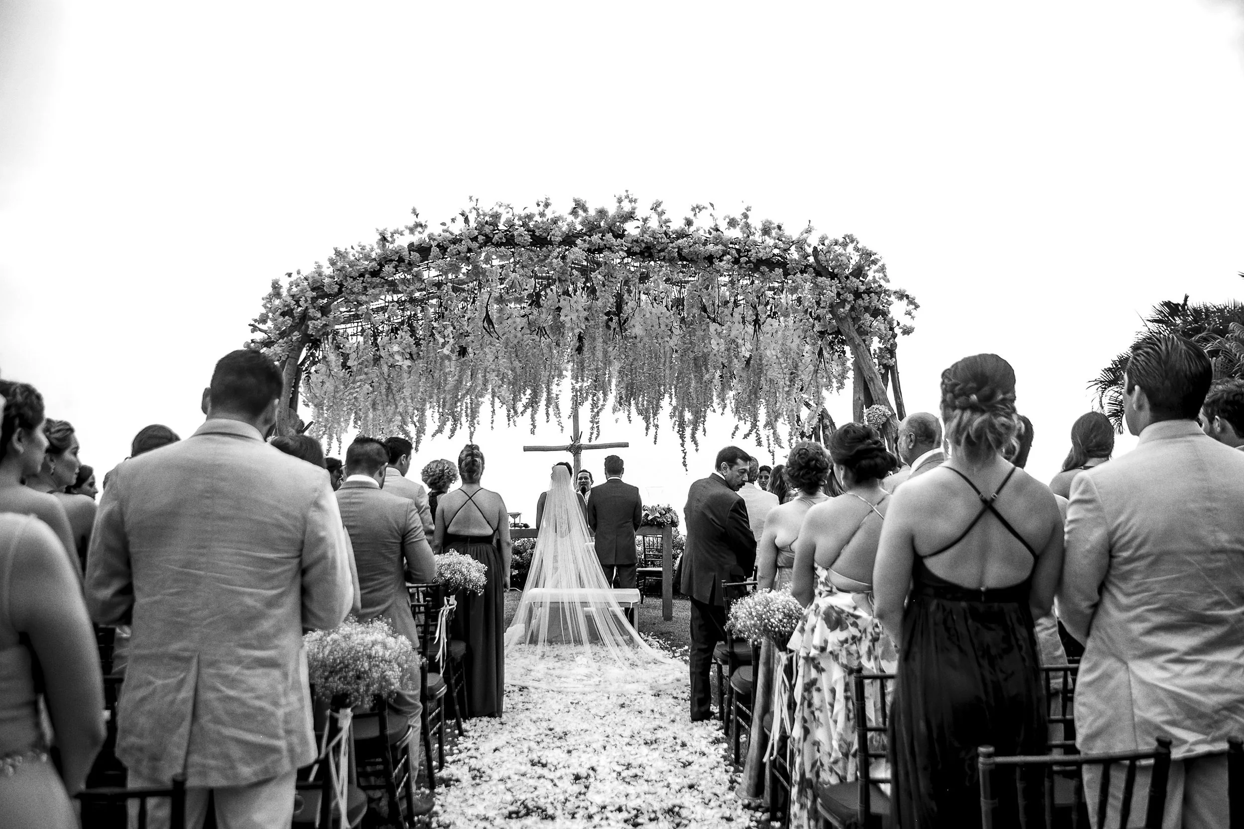 Fotografía en blanco y negro de ceremonia vista desde atrás, invitados alineados y altar floral al fondo.