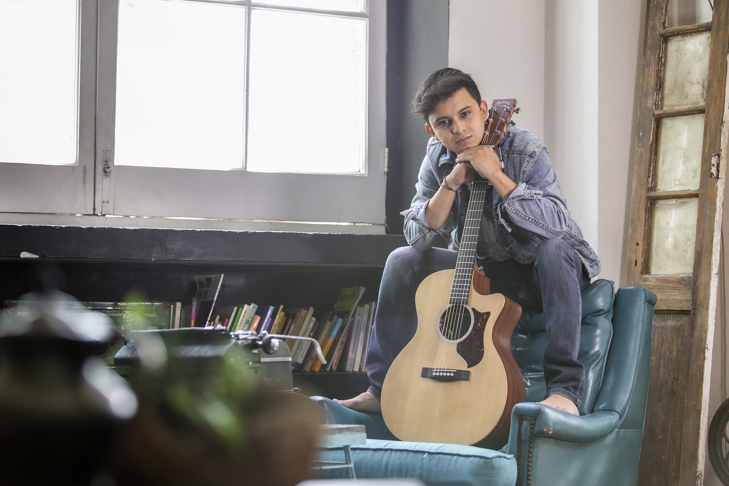 Retrato de hombre en interior con guitarra, sentado y mirada directa, ambiente doméstico.