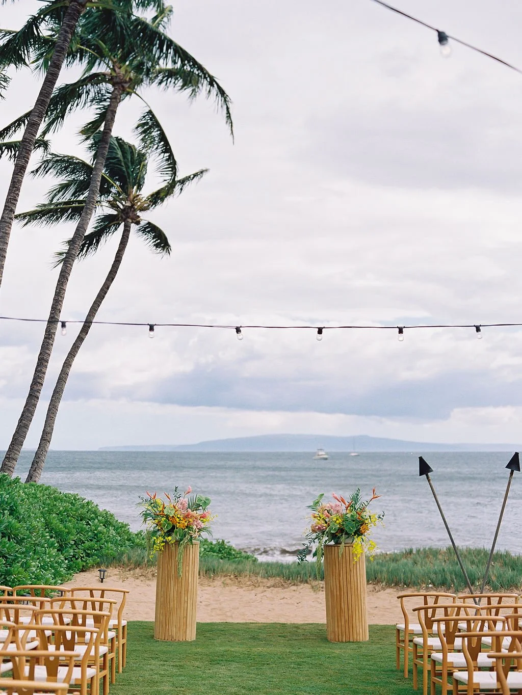 Oceanfront wedding ceremony aisle with wooden pedestals, tropical flowers and wishbone chairs in Maui
