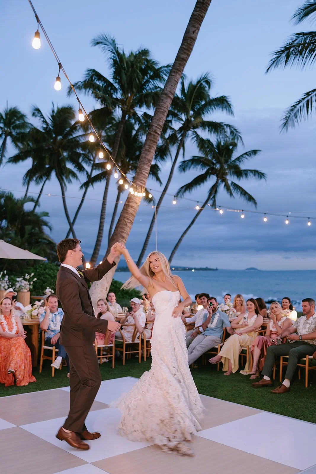 Couple sharing a first dance on a checkered dance floor at dusk beneath palm trees and string lights in Maui