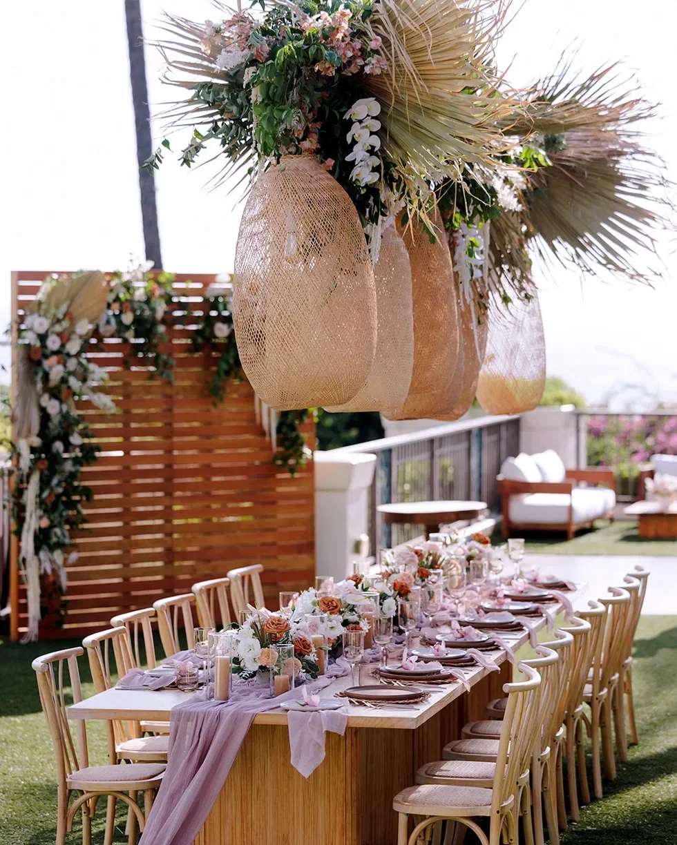Long wooden wedding table with Colette chairs, a mauve linen runner and woven pendant lanterns hanging overhead in Maui