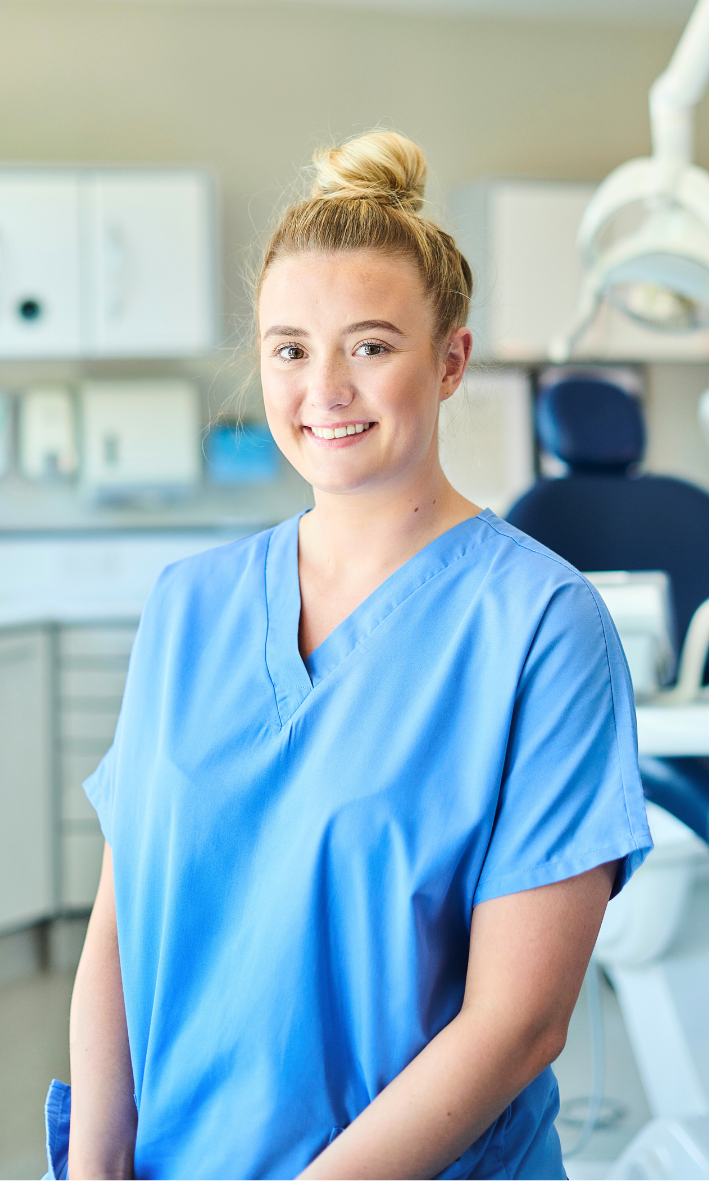 A young woman in medical scrubs standing in a clinical setting, smiling at the camera.