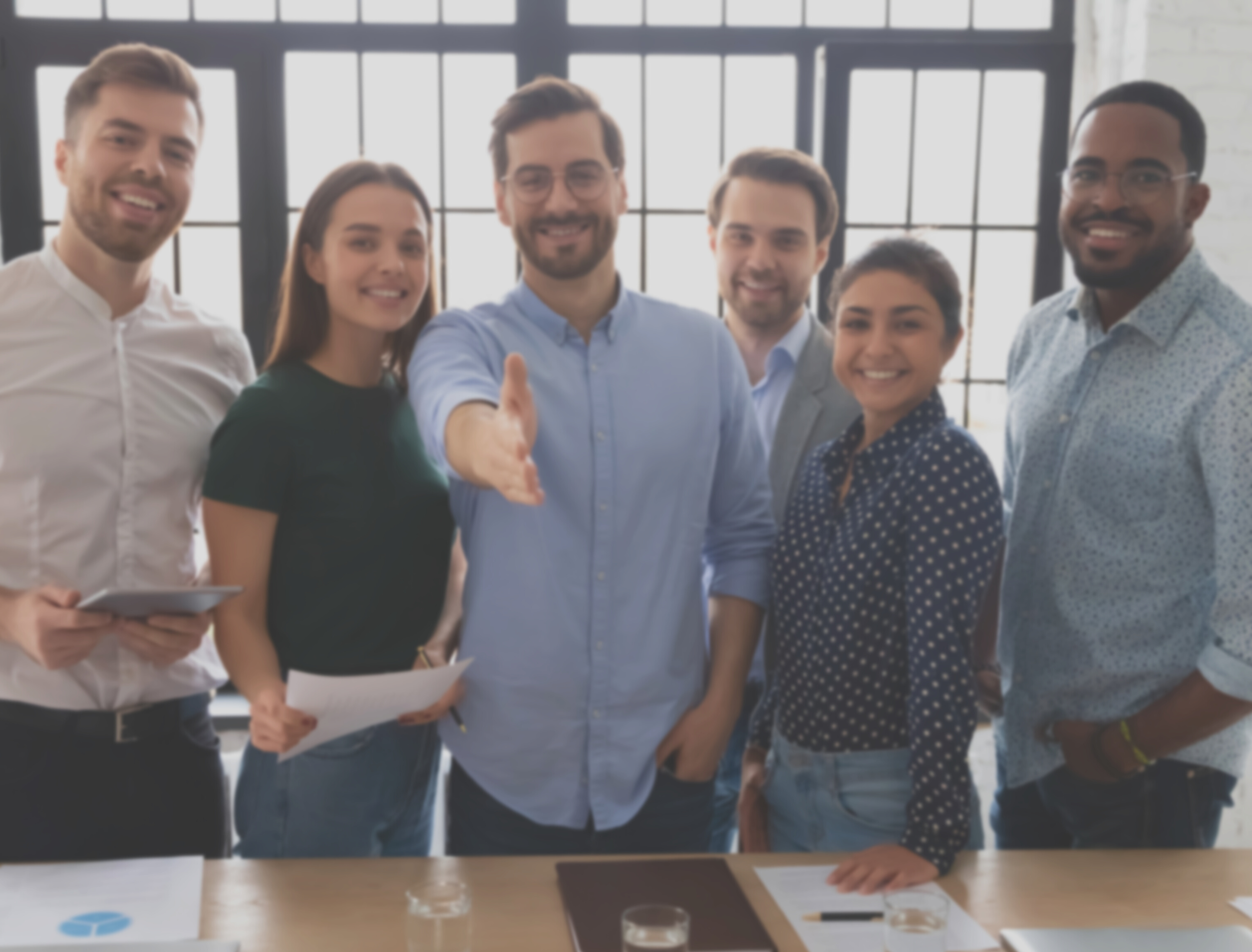 A group of seven diverse professionals standing together in a modern office, smiling, with large windows in the background. They are casually dressed, and some are holding papers or a tablet.