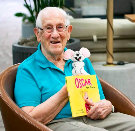 An elderly man smiling and sitting on a chair, holding a colorful children's book titled 'Oscar the Mouse' and a plush mouse toy.