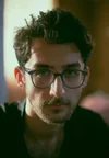 Close-up of a young man with glasses, dark hair, and a beard, looking directly at the camera, indoors with soft lighting.