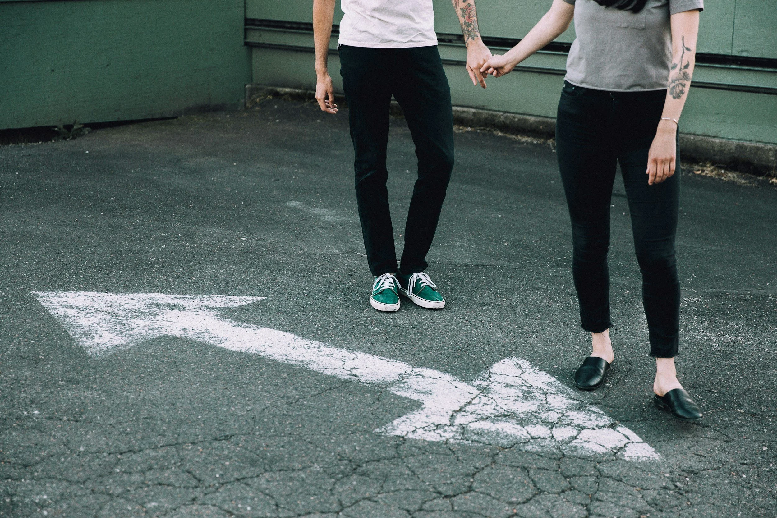 Two people on a street holding pinky fingers together in a "pinky promise" gesture, standing near crosswalk markings — one in green sneakers, the other in black flats