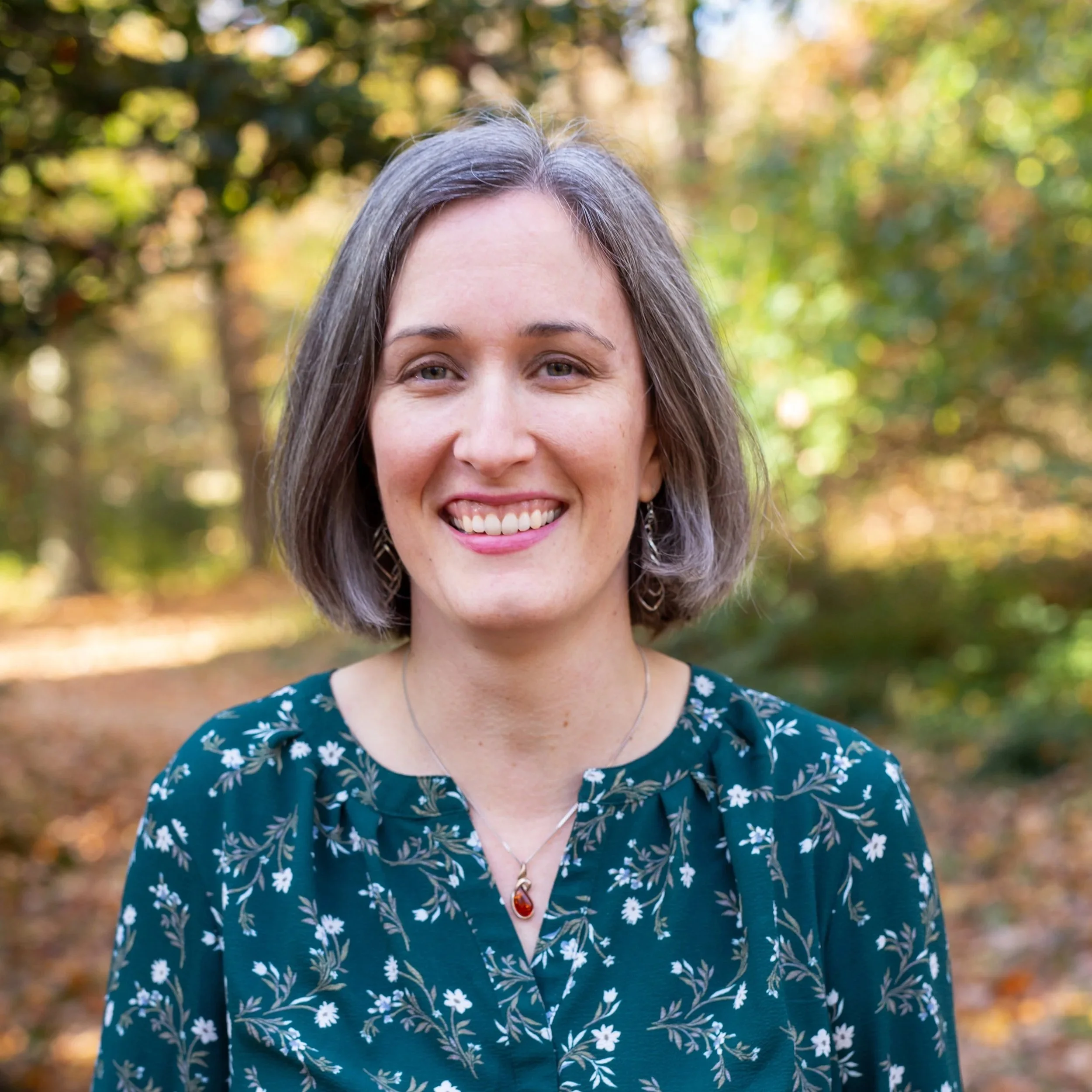 Rachel with shoulder-length gray hair smiling outdoors, with autumn leaves and trees in the background, wearing a dark green floral blouse and a necklace.