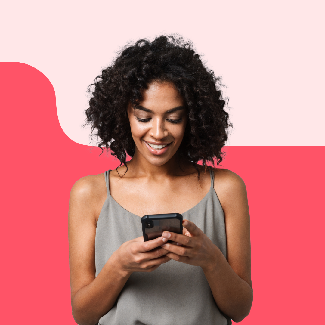 A young woman with curly black hair, wearing a gray sleeveless top, is looking at her phone and smiling against a pink and red background.