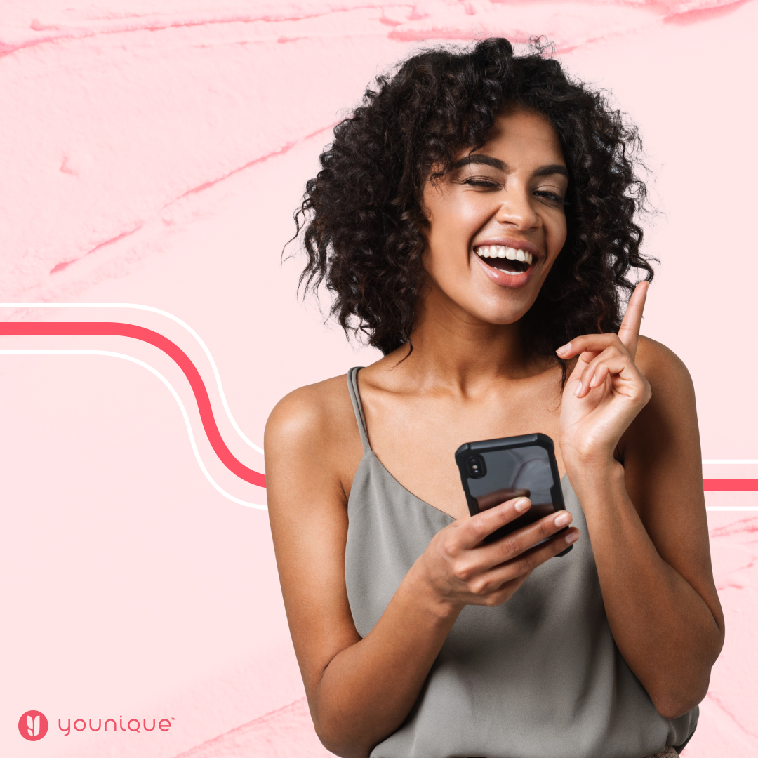 Smiling woman with curly hair holding a phone and pointing upward, against a pink background with a graphic design.
