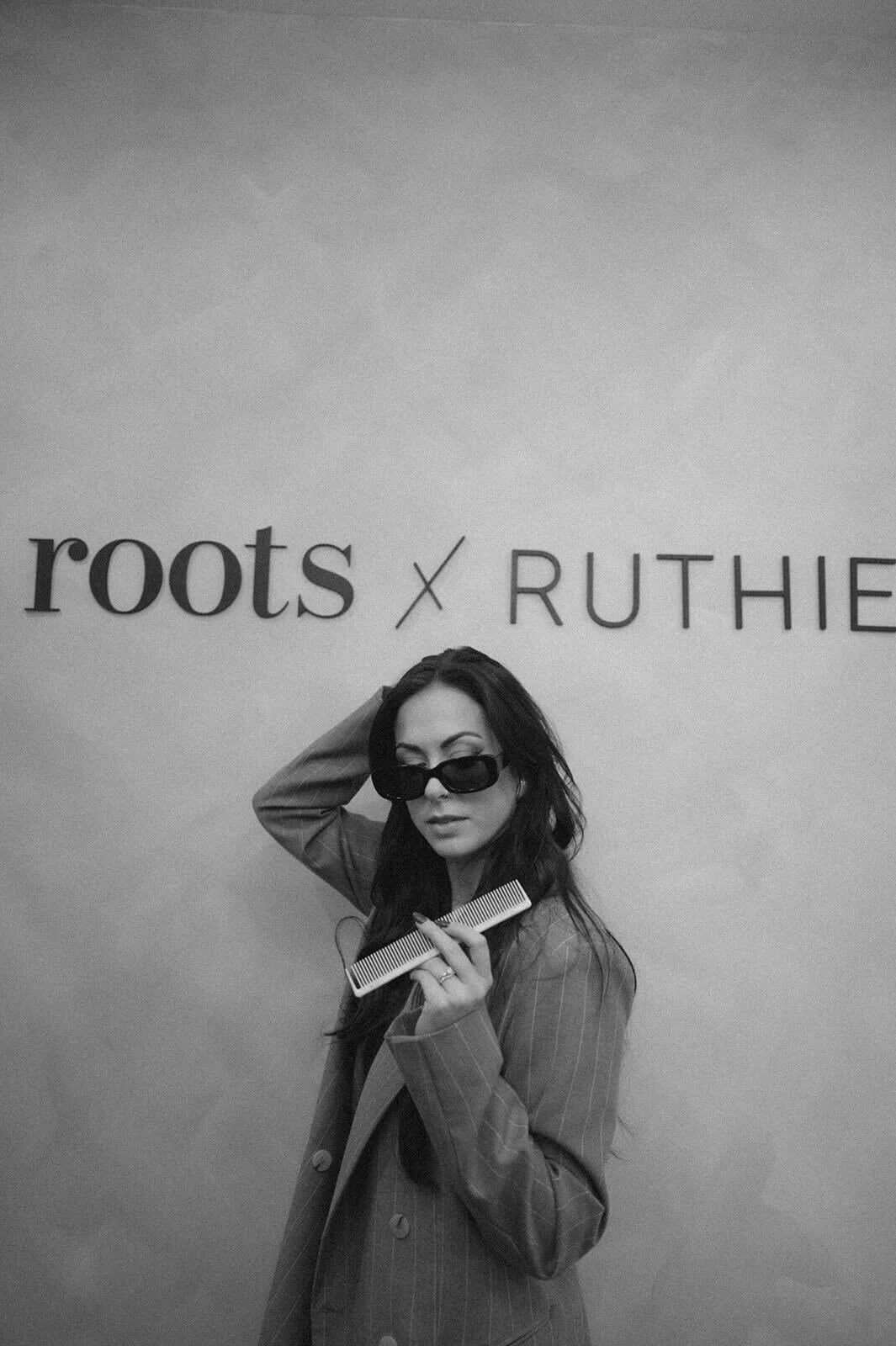 A woman with dark hair wearing sunglasses and a pinstripe blazer holding a comb, posing in front of a wall with the words 'roots x RUTHIE' in black lettering.