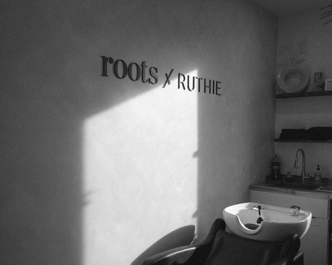 A black and white photo of a salon interior with a wall sign that reads "roots x Ruthie" and a washbasin chair in the foreground.
