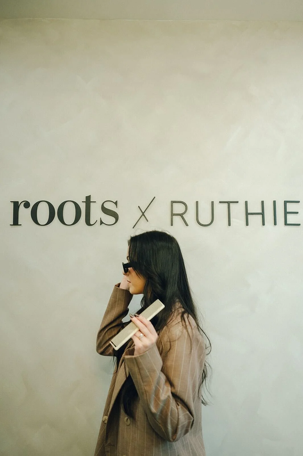 A woman with long dark hair wearing a brown plaid blazer and black glasses, holding a hairbrush, standing in front of a wall with the words 'roots' and 'RUTHE' on it.