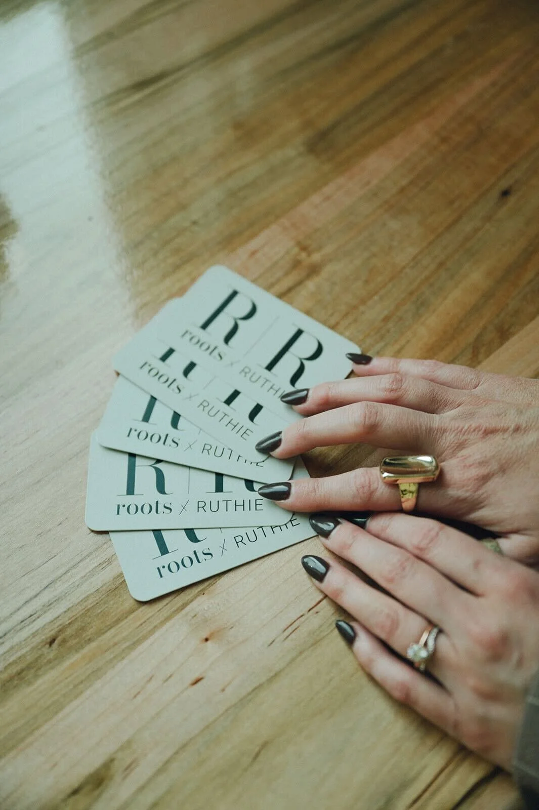 A hand with dark nail polish and rings resting on a wooden surface near several cards that read "roots x RUTHIE."