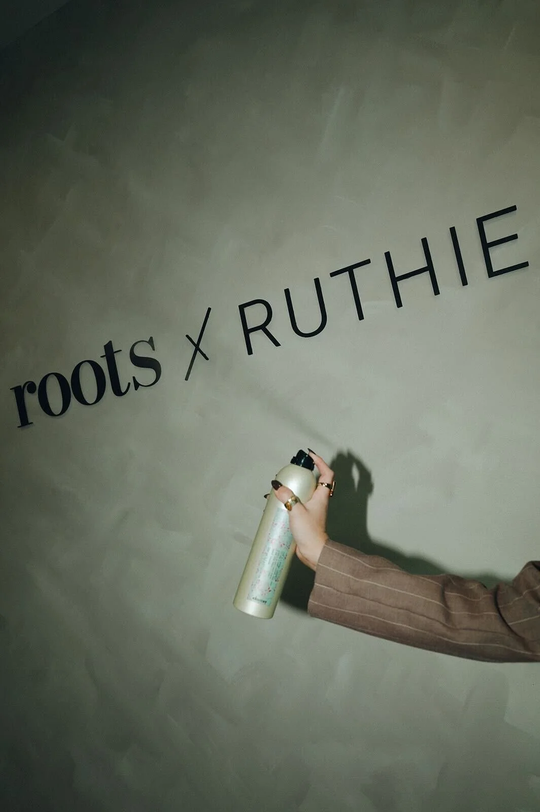 Person holding a spray can near a wall with the words 'roots x RUTHIE' printed on it.