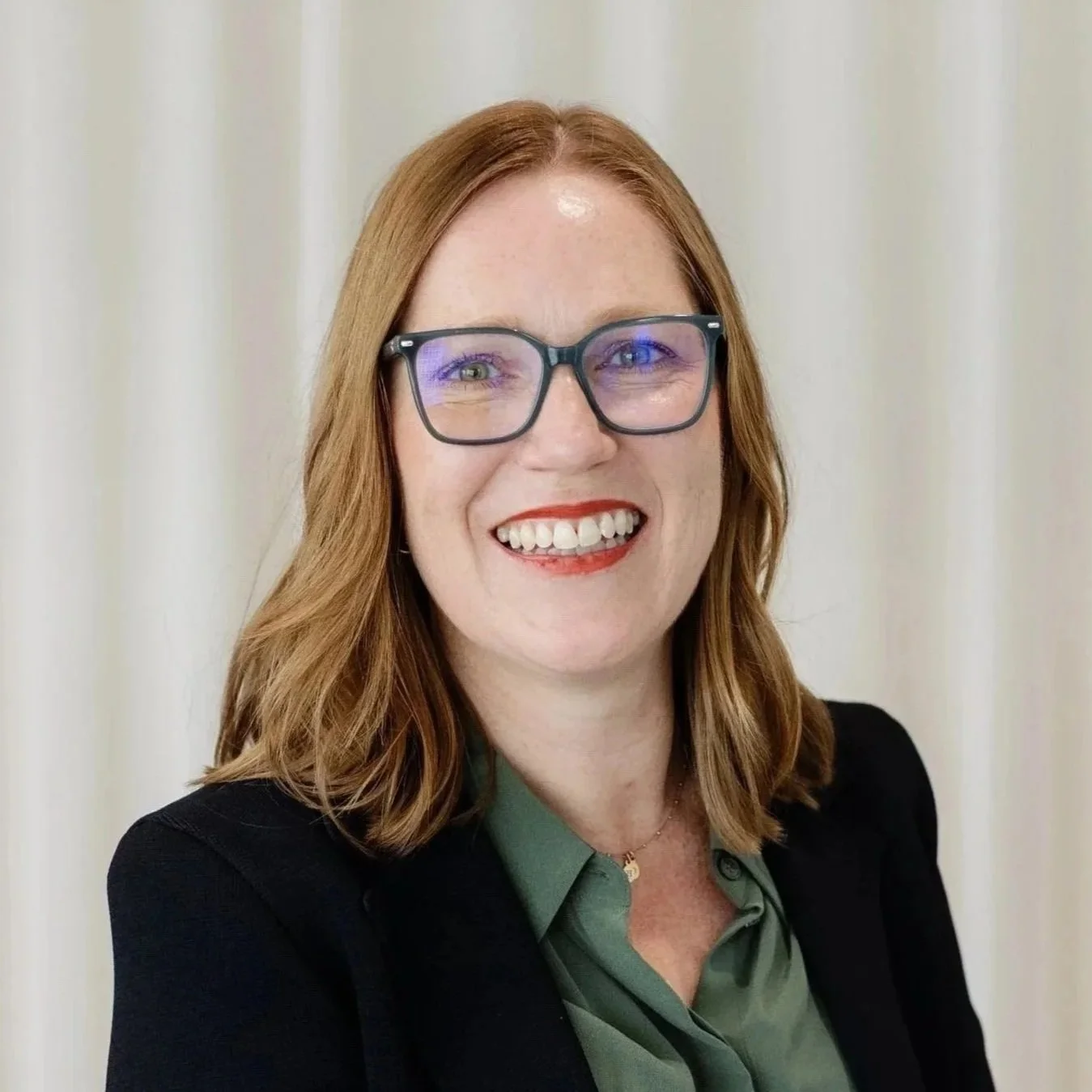 A smiling woman with red hair, glasses, wearing a dark blazer over a green blouse, standing in front of a light-colored curtain.