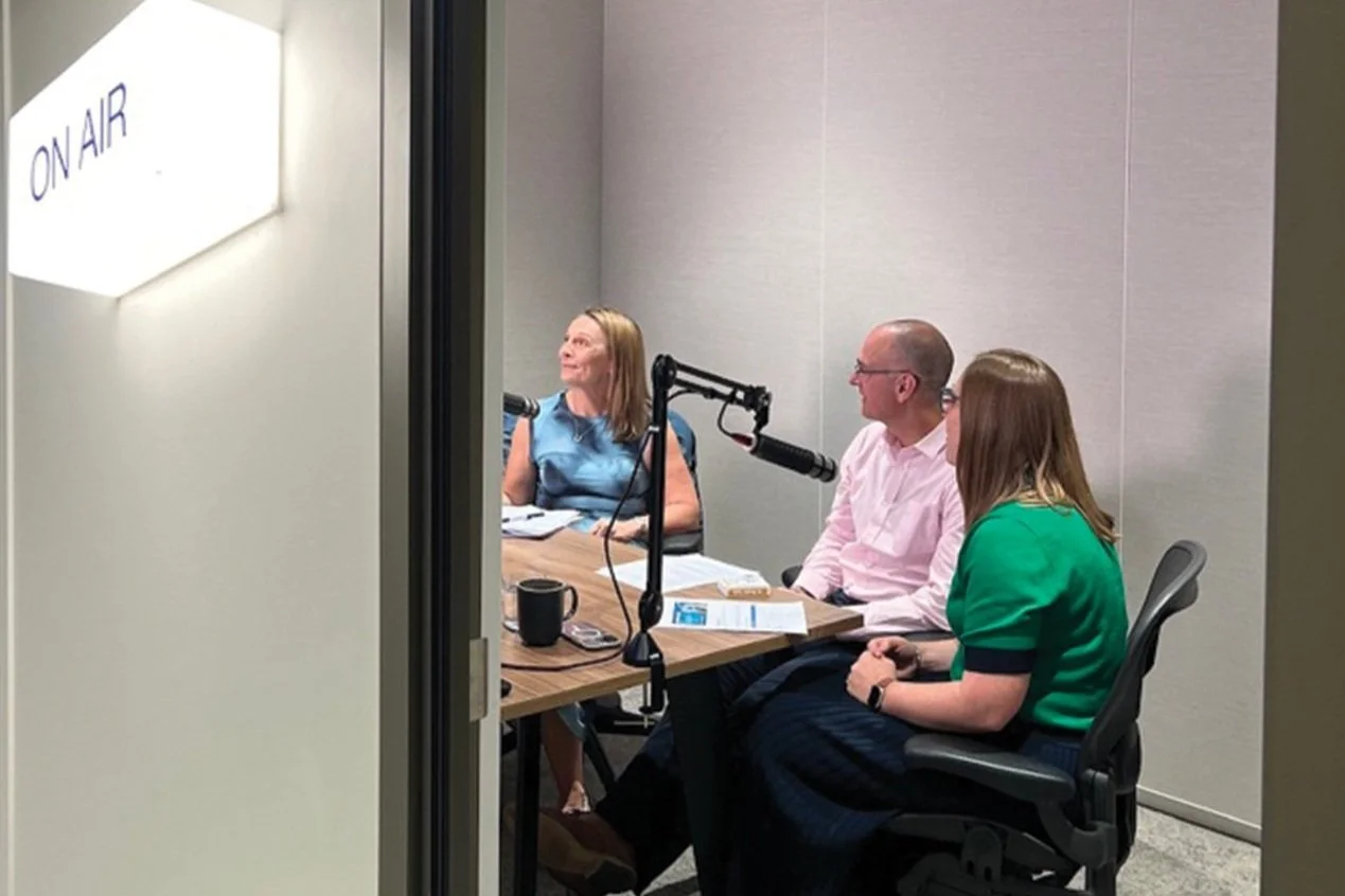 Three people in a recording studio with a microphone, seen through a doorway with an 'On Air' sign. One woman and two men sit around a table with papers and cups, engaged in conversation.