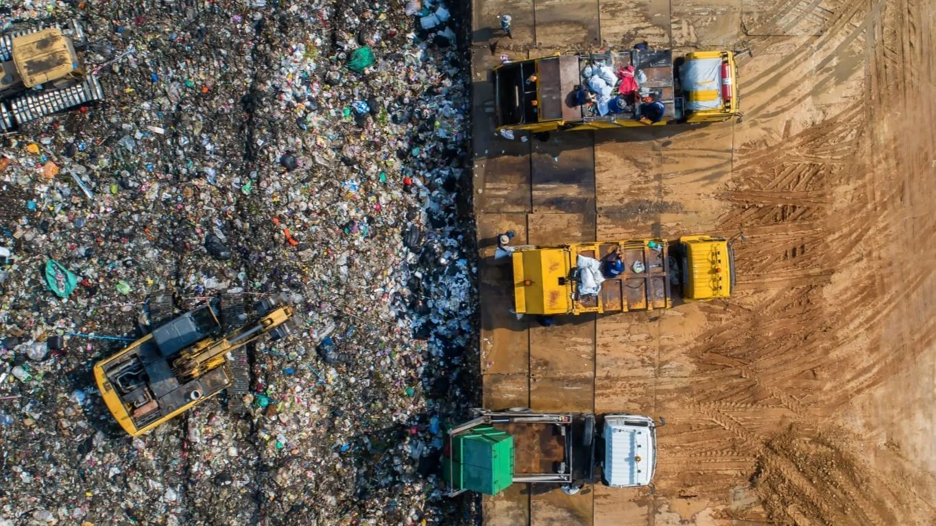 Aerial view of a garbage dump with multiple yellow trucks working beside a pile of mixed waste, cardboard, and debris.