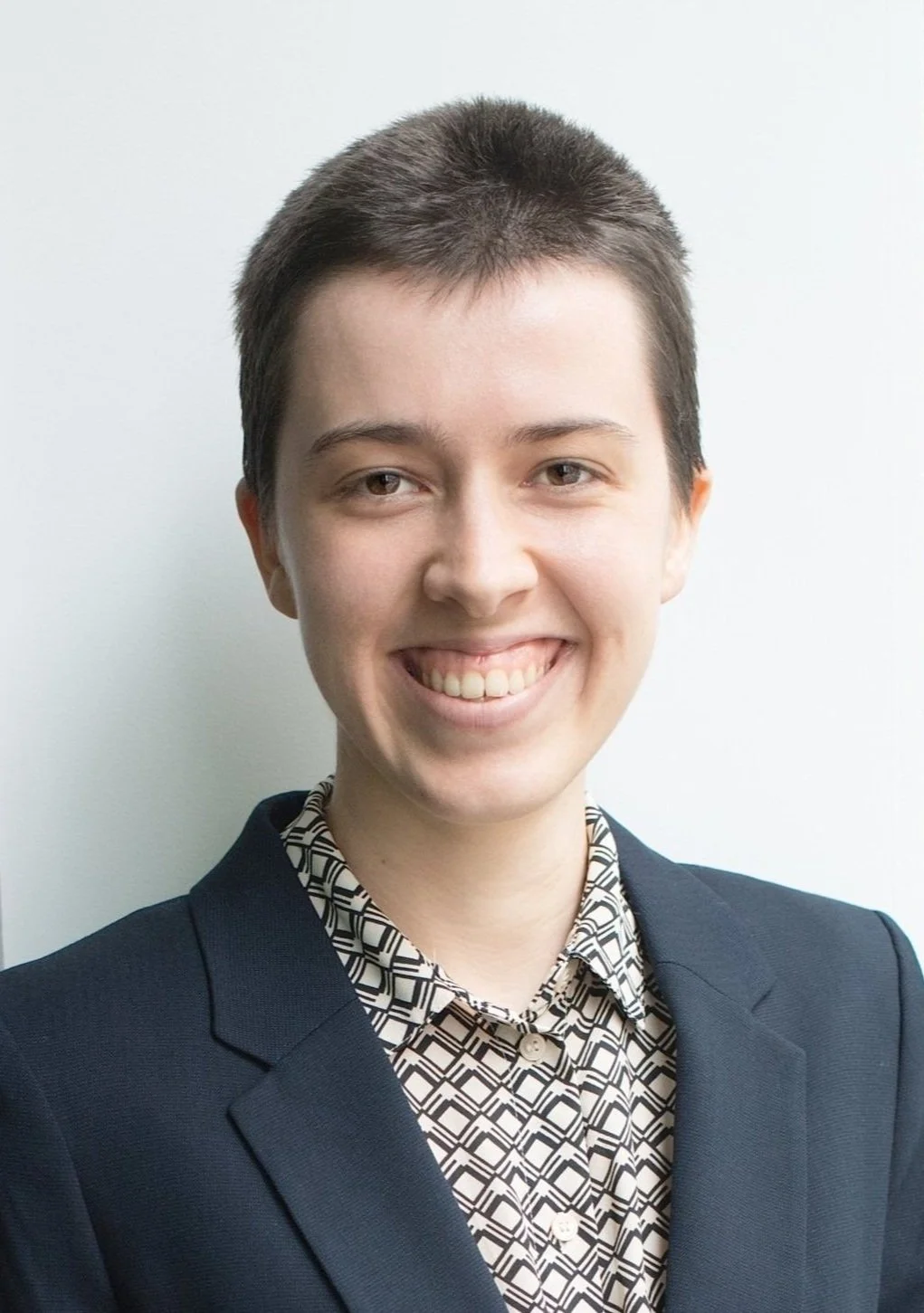 A young woman with short dark hair wearing a dark blazer and a patterned shirt, smiling against a plain background.