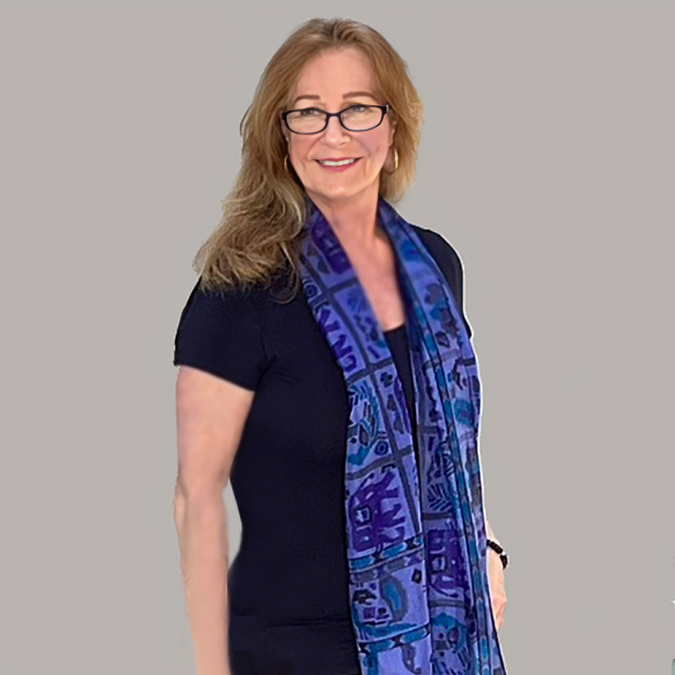 A woman with light skin, long wavy red hair, wearing glasses, a black top, and a blue patterned scarf, smiling and standing against a plain gray background.