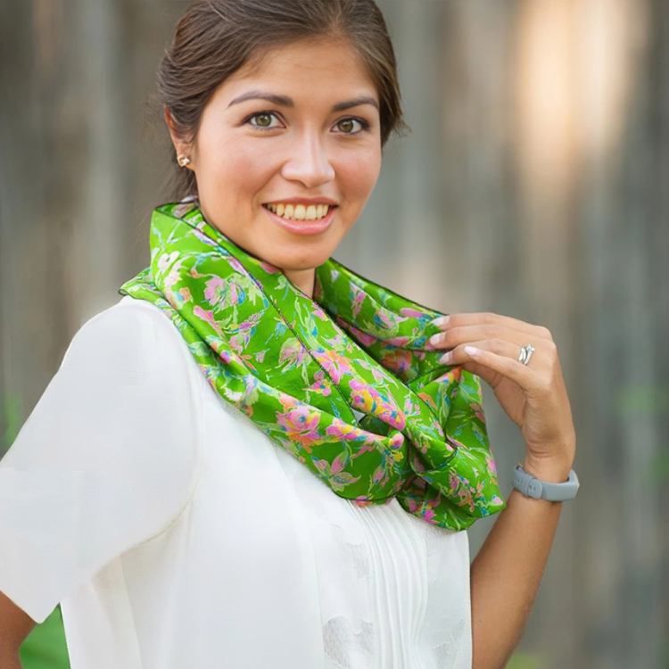 A woman smiling outdoors wearing a white top and a green floral scarf, holding the scarf with one hand.