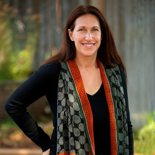 A smiling woman with long brown hair standing outdoors near a wooden fence, wearing a black shirt and a patterned scarf with leaf motifs and red borders.