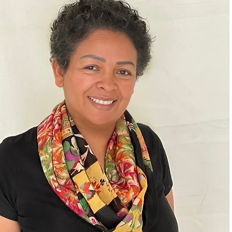 A woman with short curly hair smiling, wearing a black top and a colorful floral scarf, against a light background.