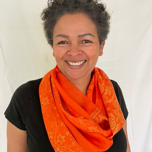 A woman with short, curly gray hair smiling, wearing a black shirt and an orange scarf, standing against a plain white background.