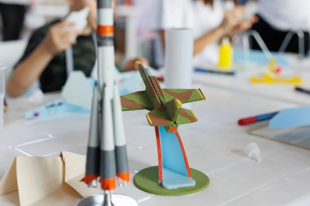 Model rocket and aircraft crafts on a workshop table, with students assembling designs in the background during a hands-on STEM activity.
