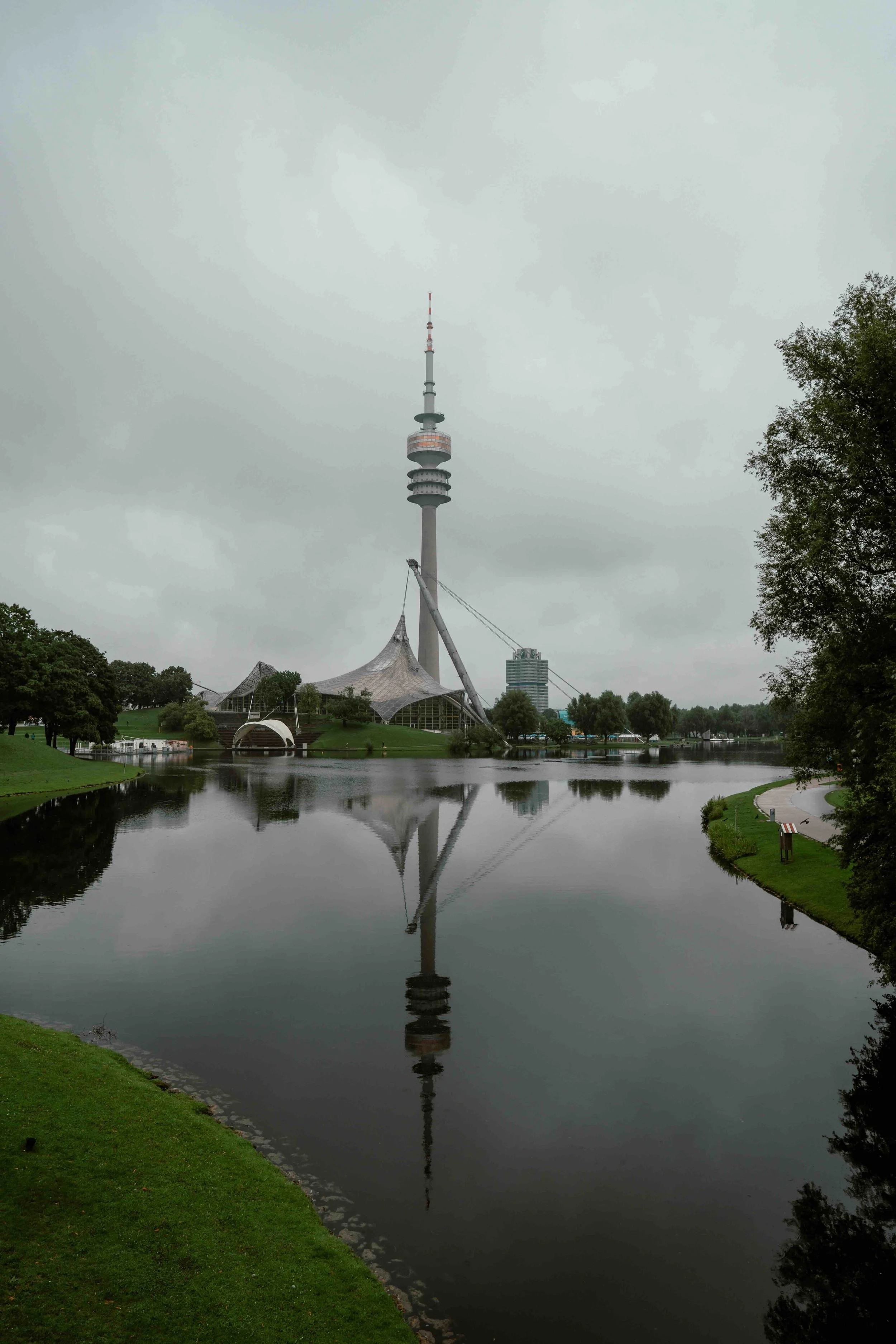 Munich, Germany | The Olympic Tower (Olympiaturm) reflected in the Olympic Lake under an overcast sky.