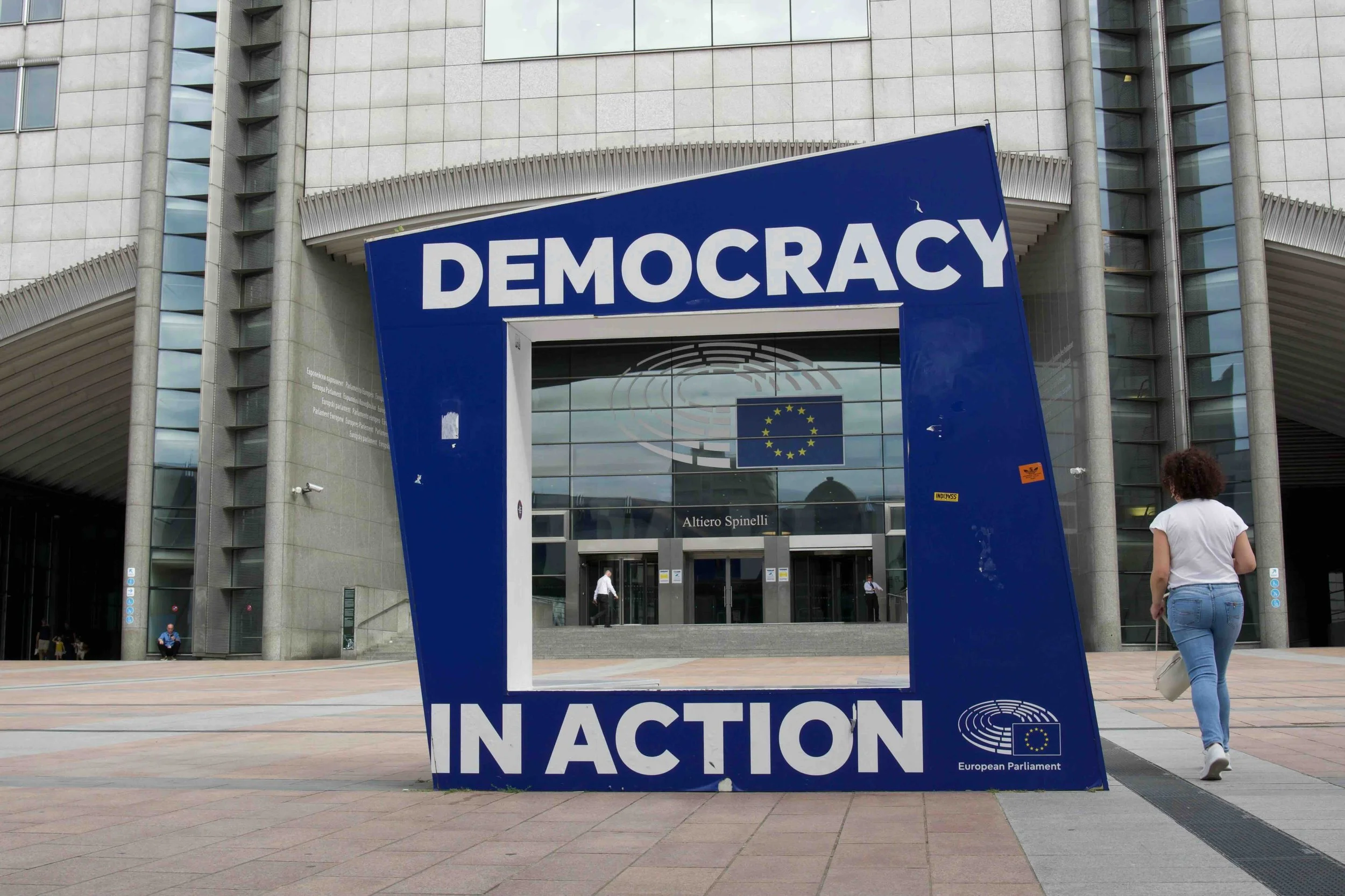 Brussels, Belgium | "Democracy in Action" sign frame outside the European Parliament's Altiero Spinelli building, with a pedestrian walking past.