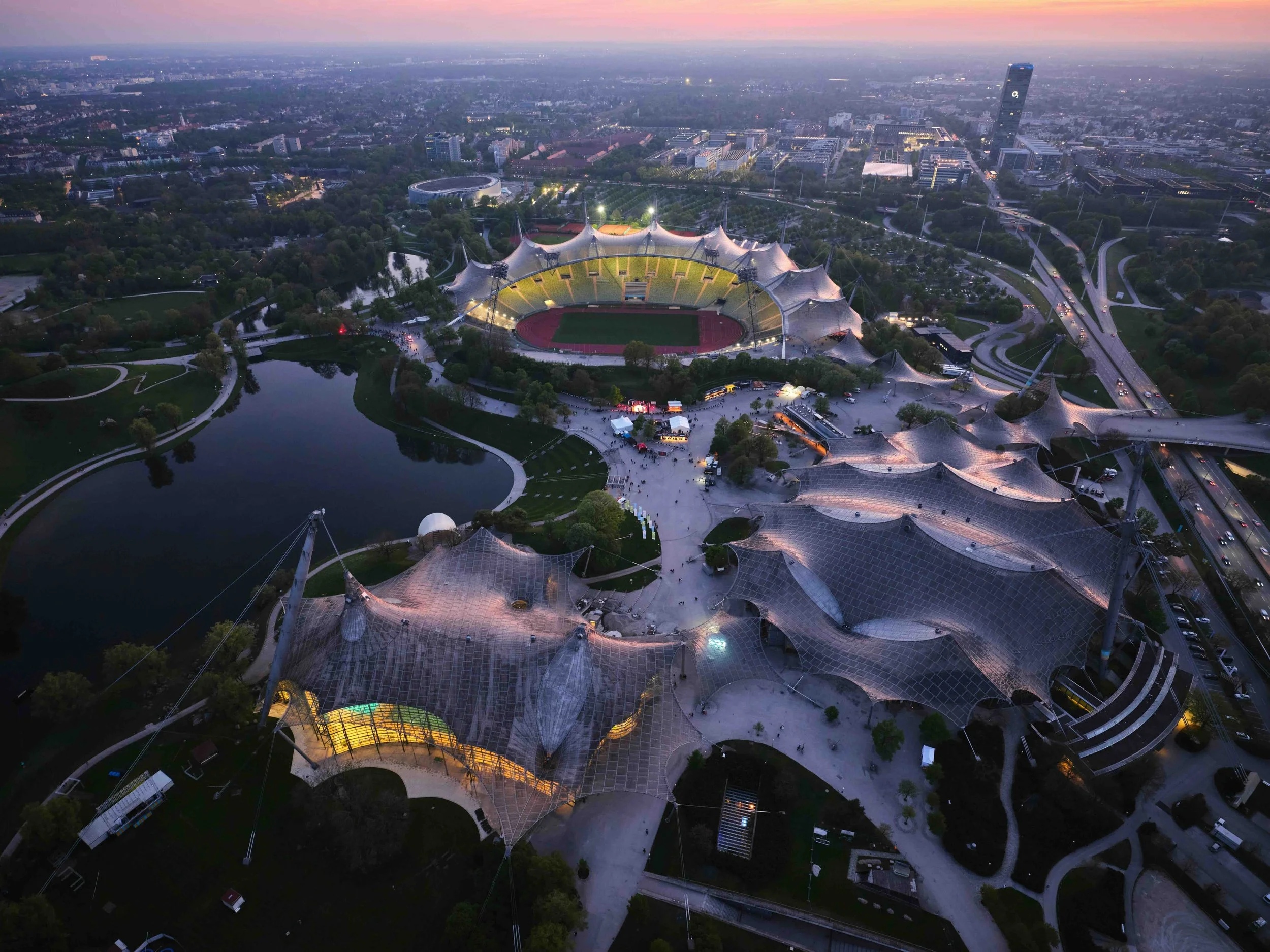 Munich, Germany | Aerial dusk view of the Olympiapark, showing the tent-like roof structures and Olympic Stadium illuminated at twilight.