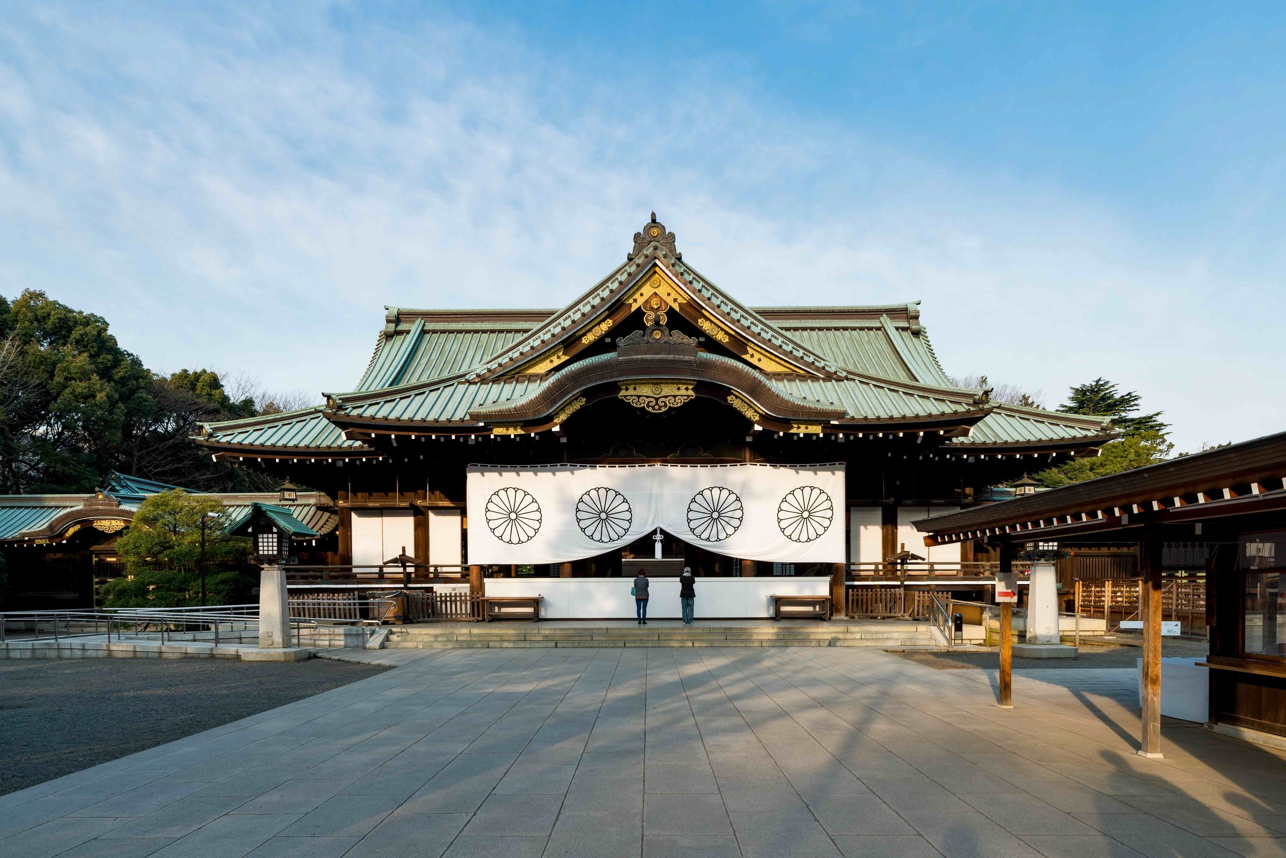 The main sanctuary (haiden) of Yasukuni Shrine in Tokyo at dawn, featuring a sweeping copper-green roof, imperial chrysanthemum crests, and a nearly empty forecourt