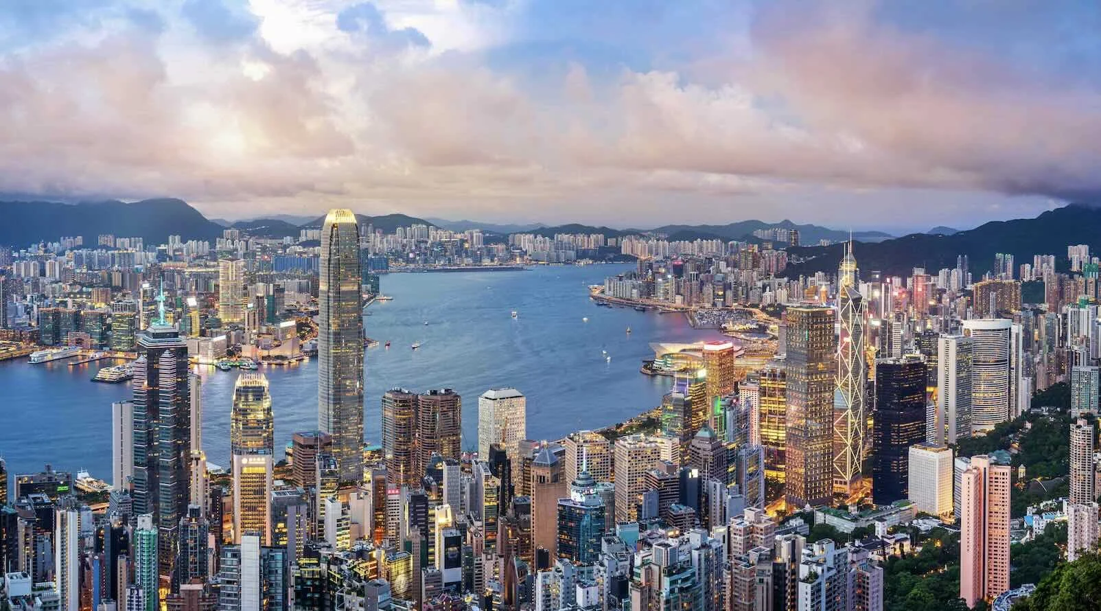 Aerial view of the Hong Kong skyline and Victoria Harbour at dusk, skyscrapers illuminated against a cloudy sky.