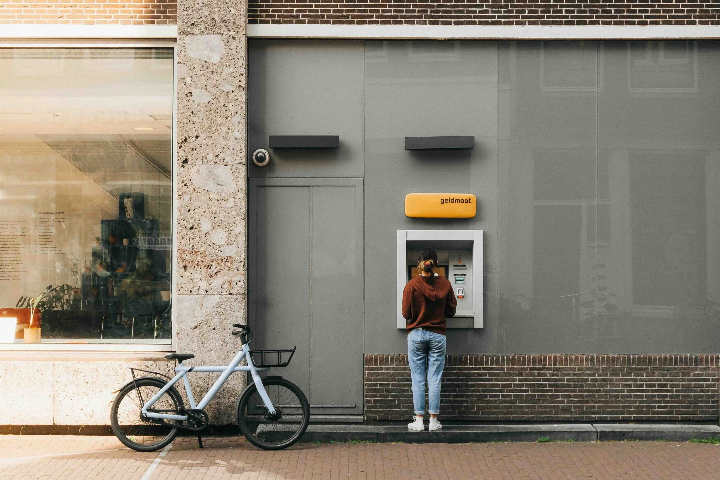 Amsterdam, Netherlands | A Geldmaat cash machine on a street, with a person withdrawing money and a bicycle parked nearby.