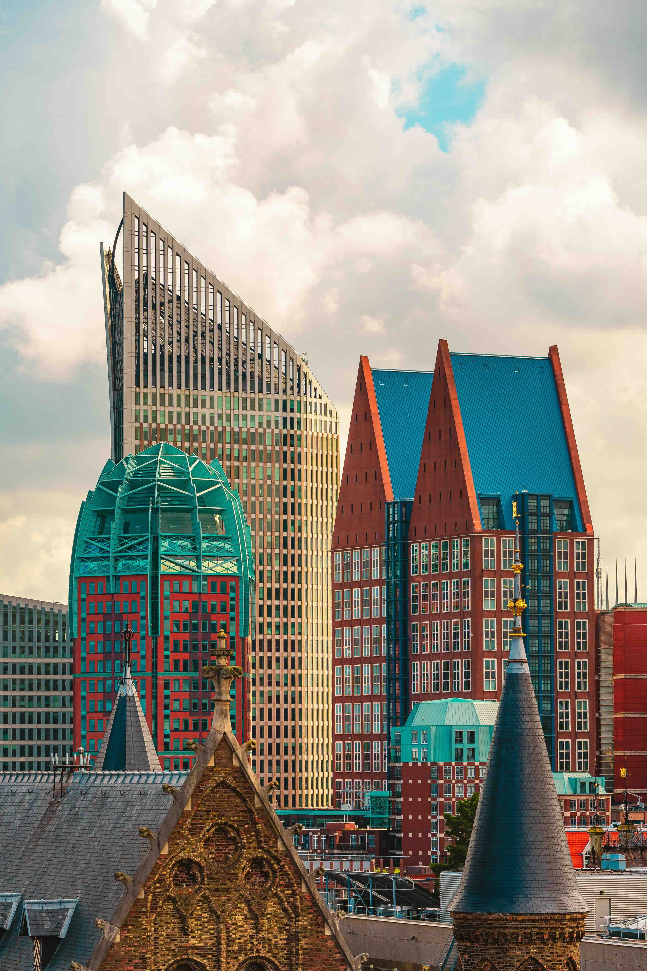 The Hague, Netherlands | Modern high-rise skyline of The Hague featuring the angular Zurichtoren and red-and-turquoise Haagse Torens, with the Gothic spires of the Grote Kerk in the foreground.