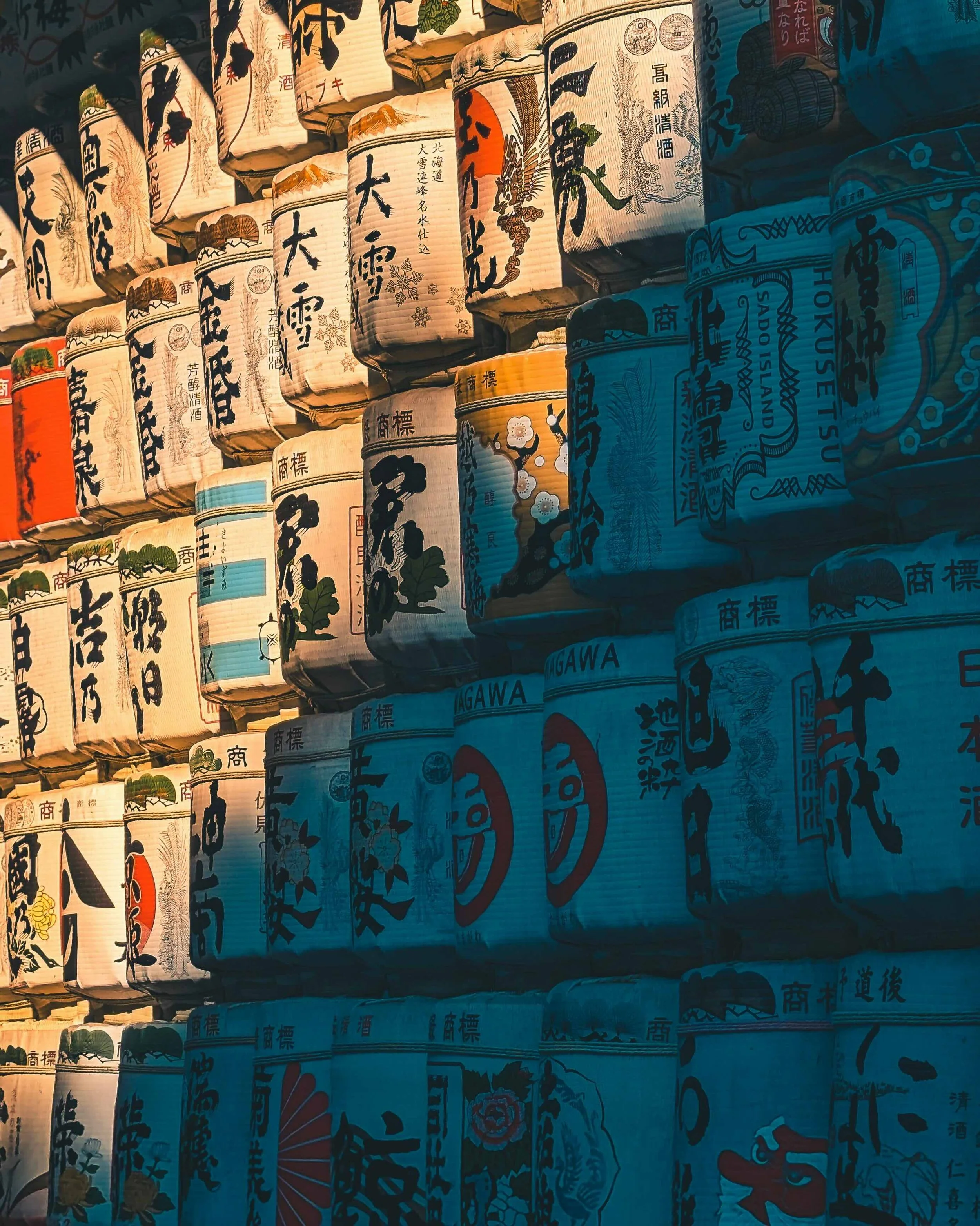 Rows of traditional Japanese sake barrels (kazaridaru) stacked at Meiji Jingu Shrine in Tokyo, decorated with kanji calligraphy and illuminated by warm raking sunlight