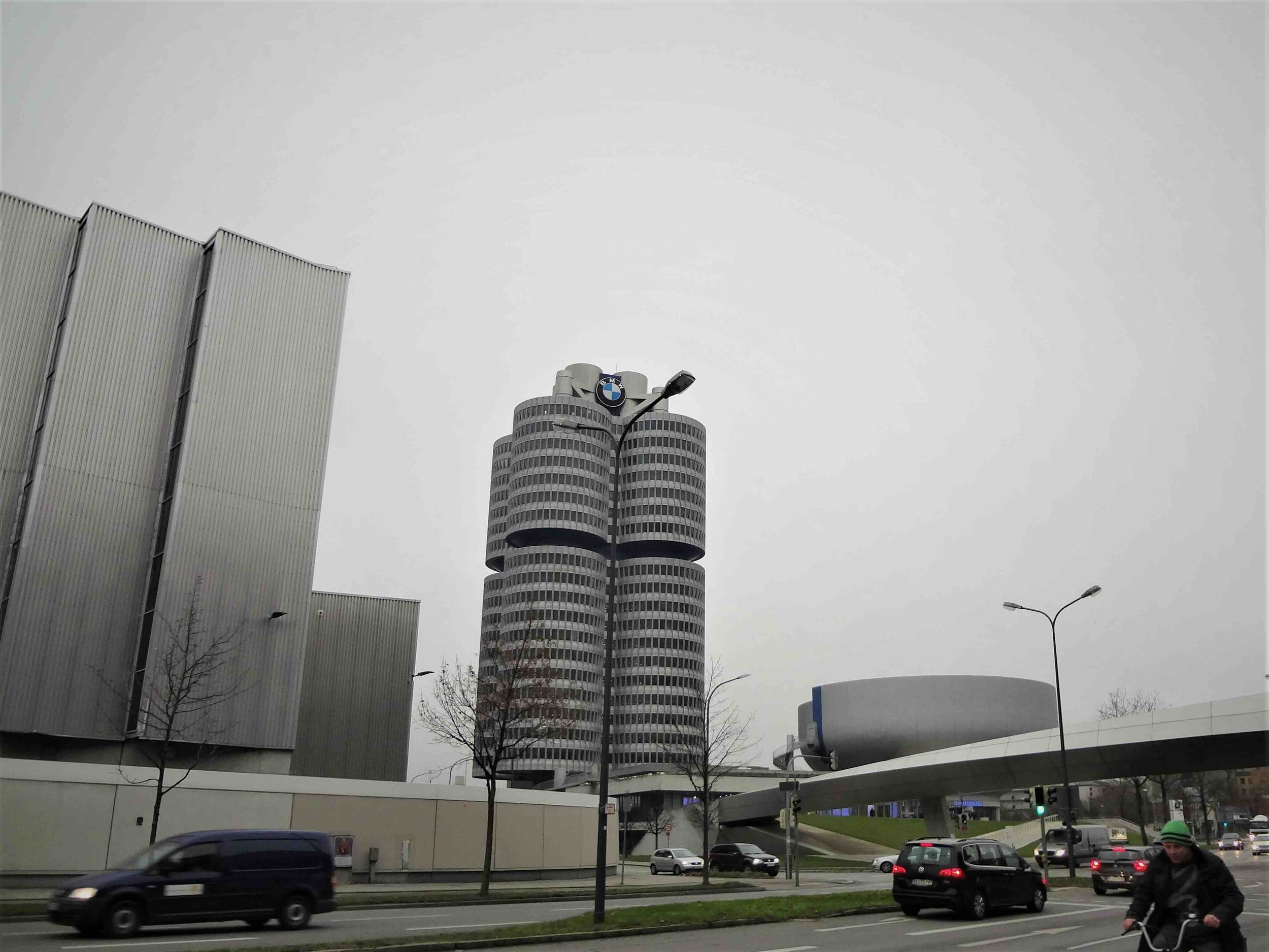 Munich, Germany | Street-level view of the BMW Headquarters tower on an overcast day, with traffic and a cyclist passing in the foreground.