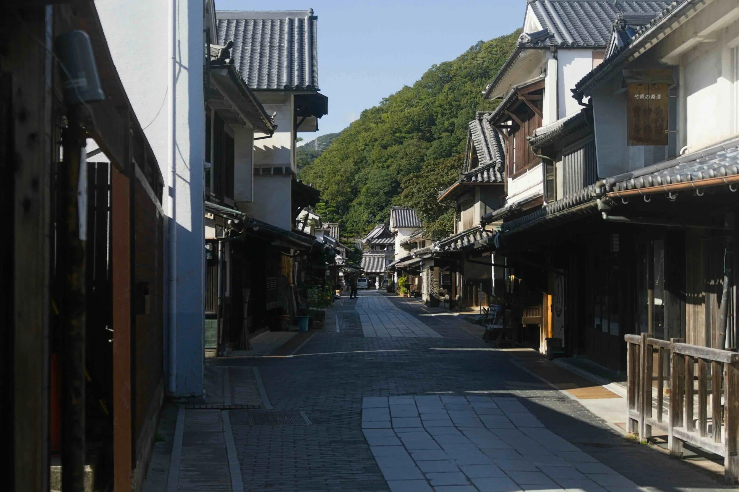 A quiet, sunlit historic merchant street in Takehara, Hiroshima Prefecture, lined with preserved Edo-period wooden townhouses and traditional tiled rooftops