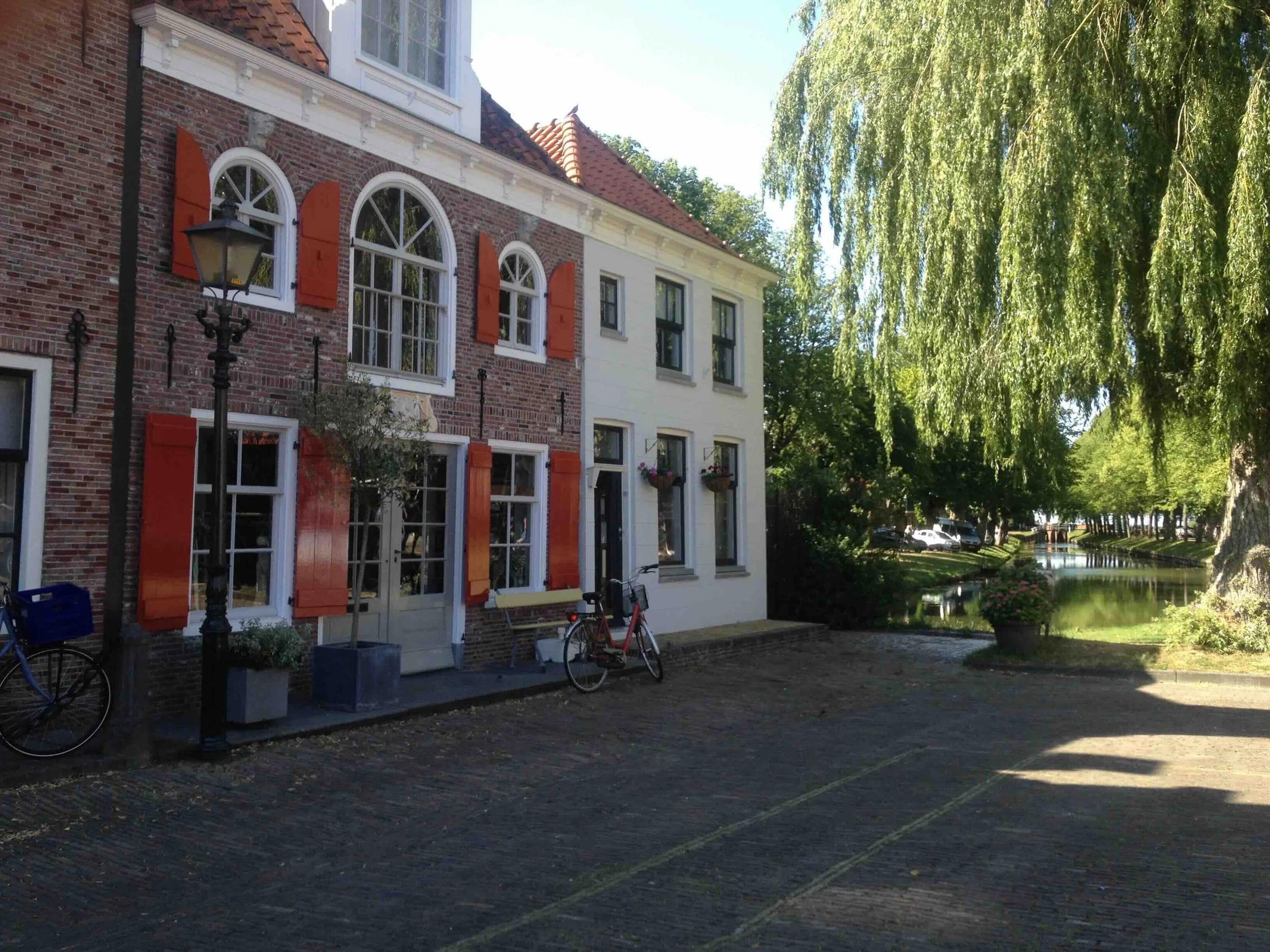 Dutch village canal street | Traditional Dutch brick townhouses with orange shutters and arched windows, bicycles parked outside, weeping willow and canal alongside a cobbled street.